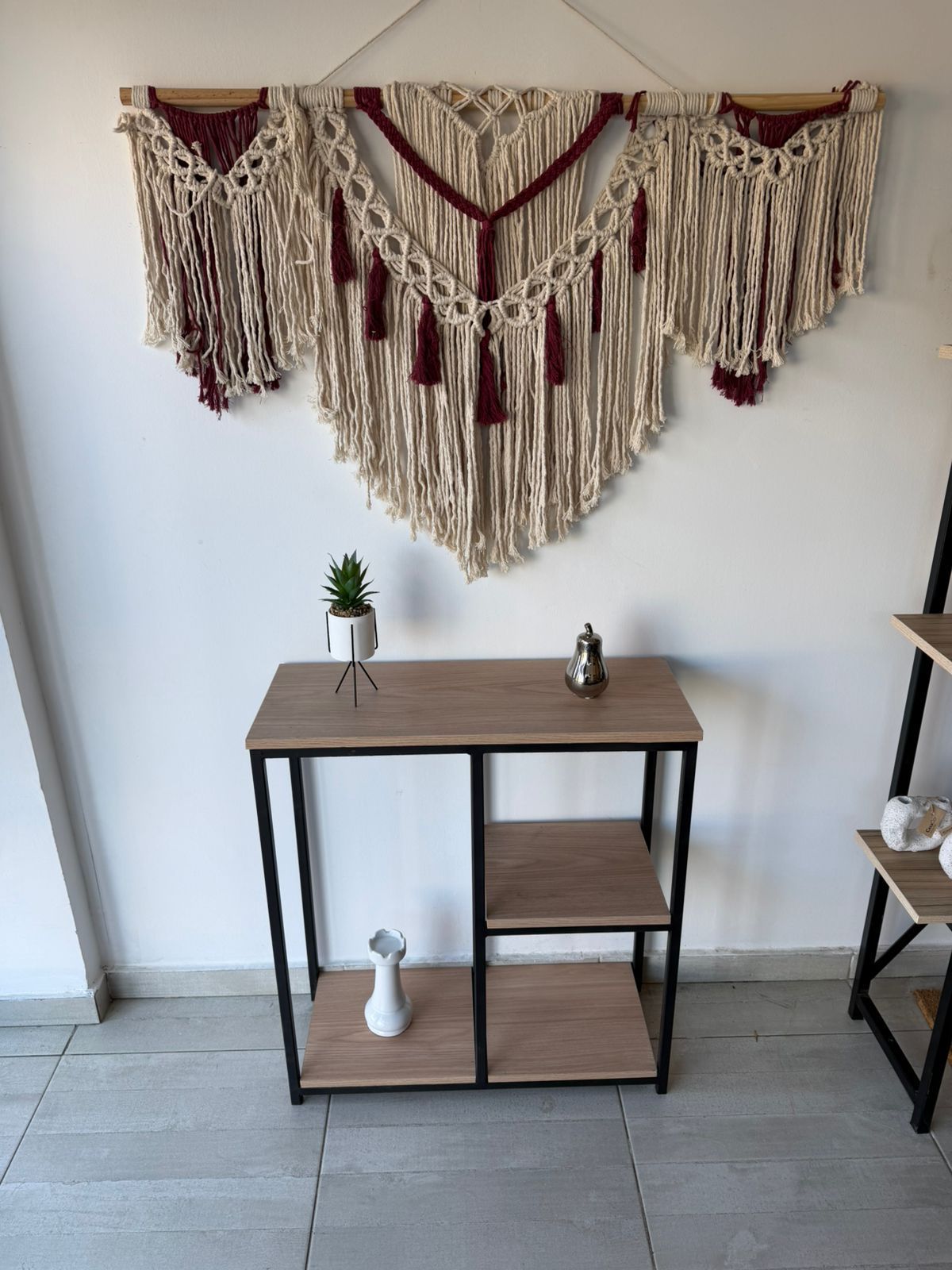 Tapiz de macramé sobre una pequeña mesa consola de madera y negra con elementos decorativos en una habitación minimalista.