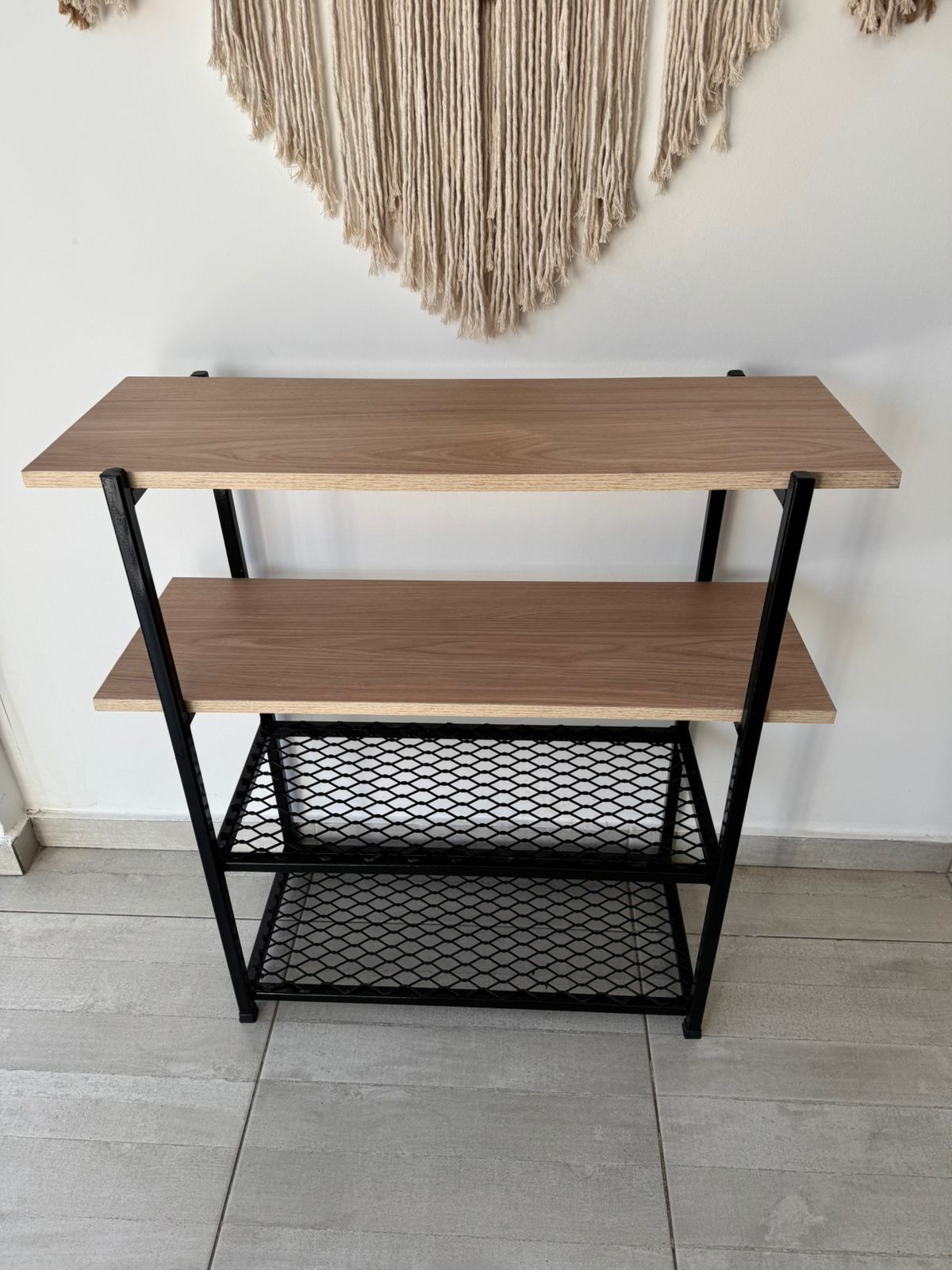 Estantería metálica negra con estantes de madera contra una pared blanca, con decoración de macramé en la parte superior.
