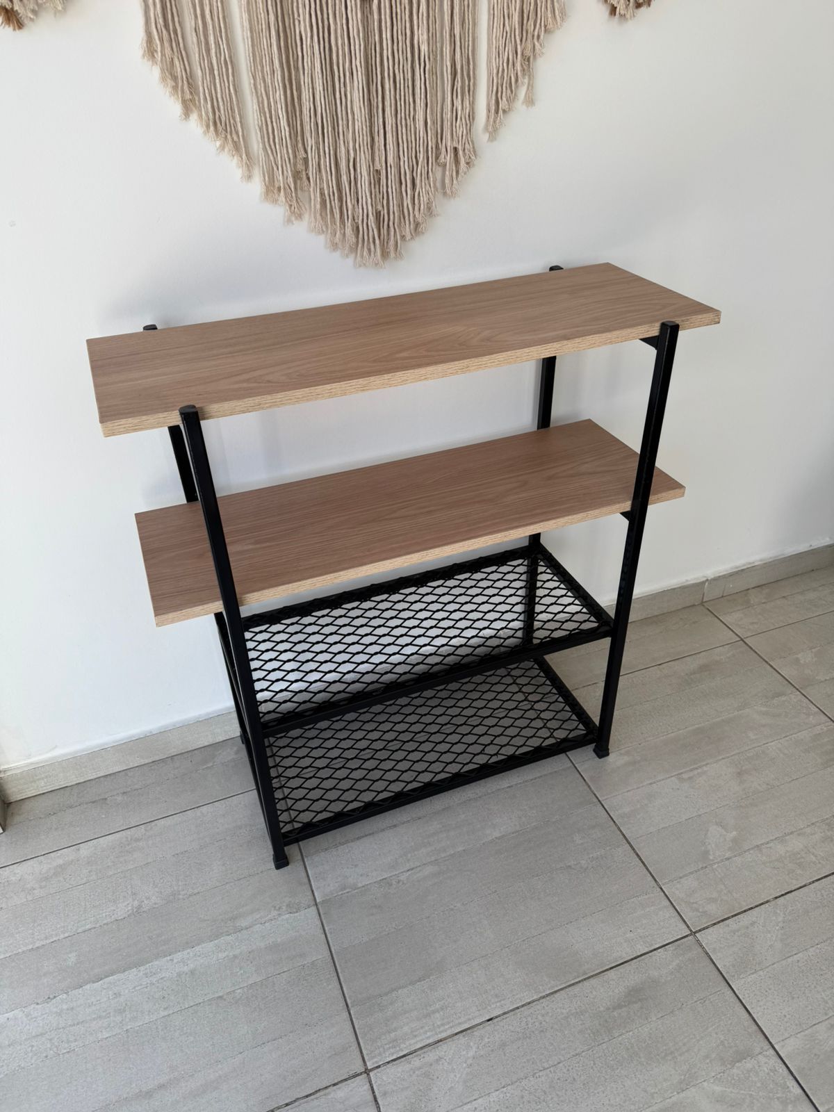Estantería metálica negra con estantes de madera contra una pared blanca, con decoración de macramé en la parte superior.