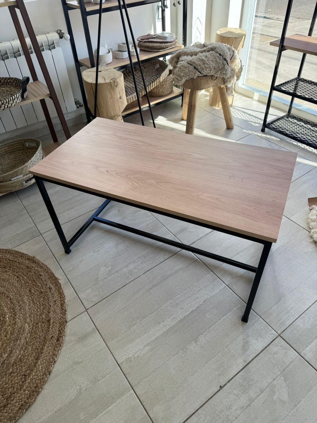 Mesa de centro rectangular con tablero de madera y patas de metal negro sobre un suelo de baldosas contra una pared blanca.