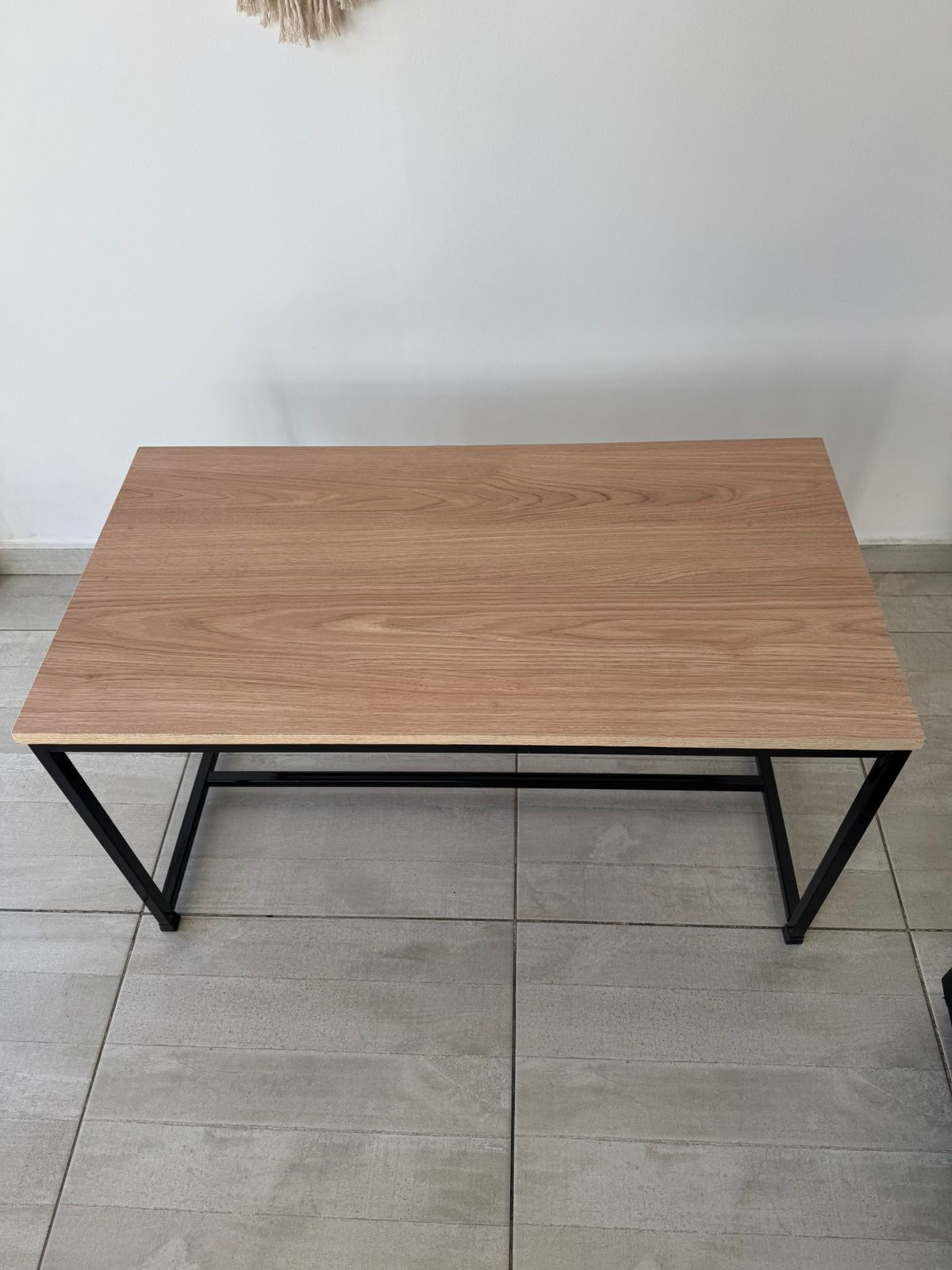 Mesa de centro rectangular con tablero de madera y patas de metal negro sobre un suelo de baldosas contra una pared blanca.