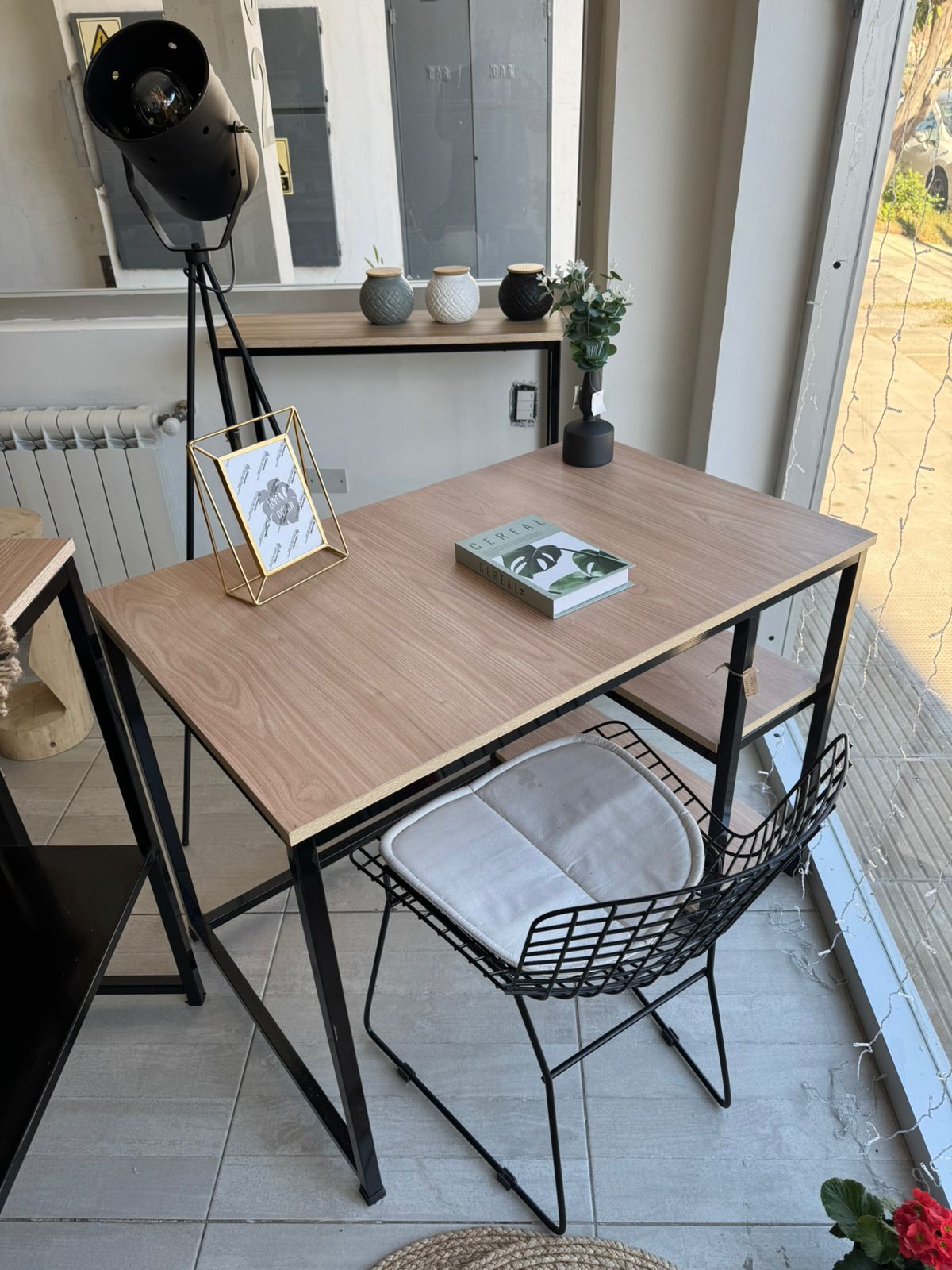 Un pequeño escritorio de madera junto a una ventana, con una silla de alambre negro.