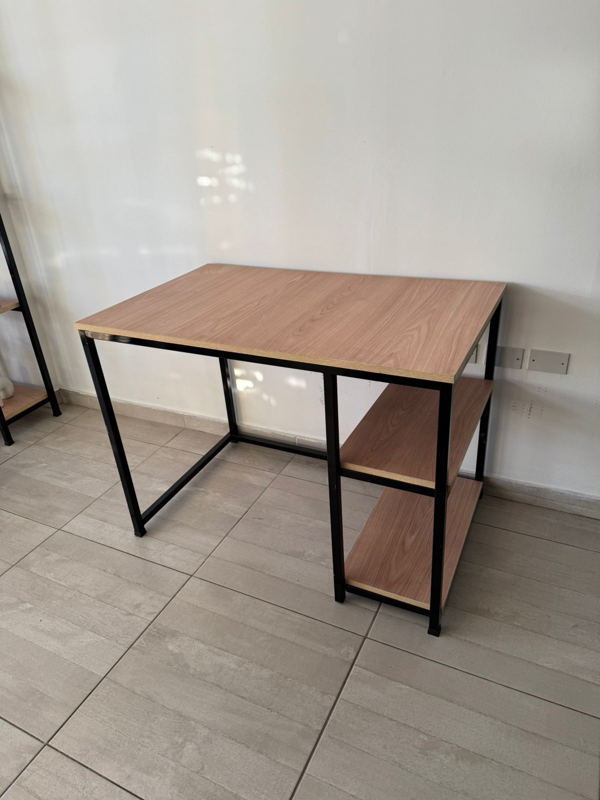 Un pequeño escritorio de madera junto a una ventana, con una silla de alambre negro.