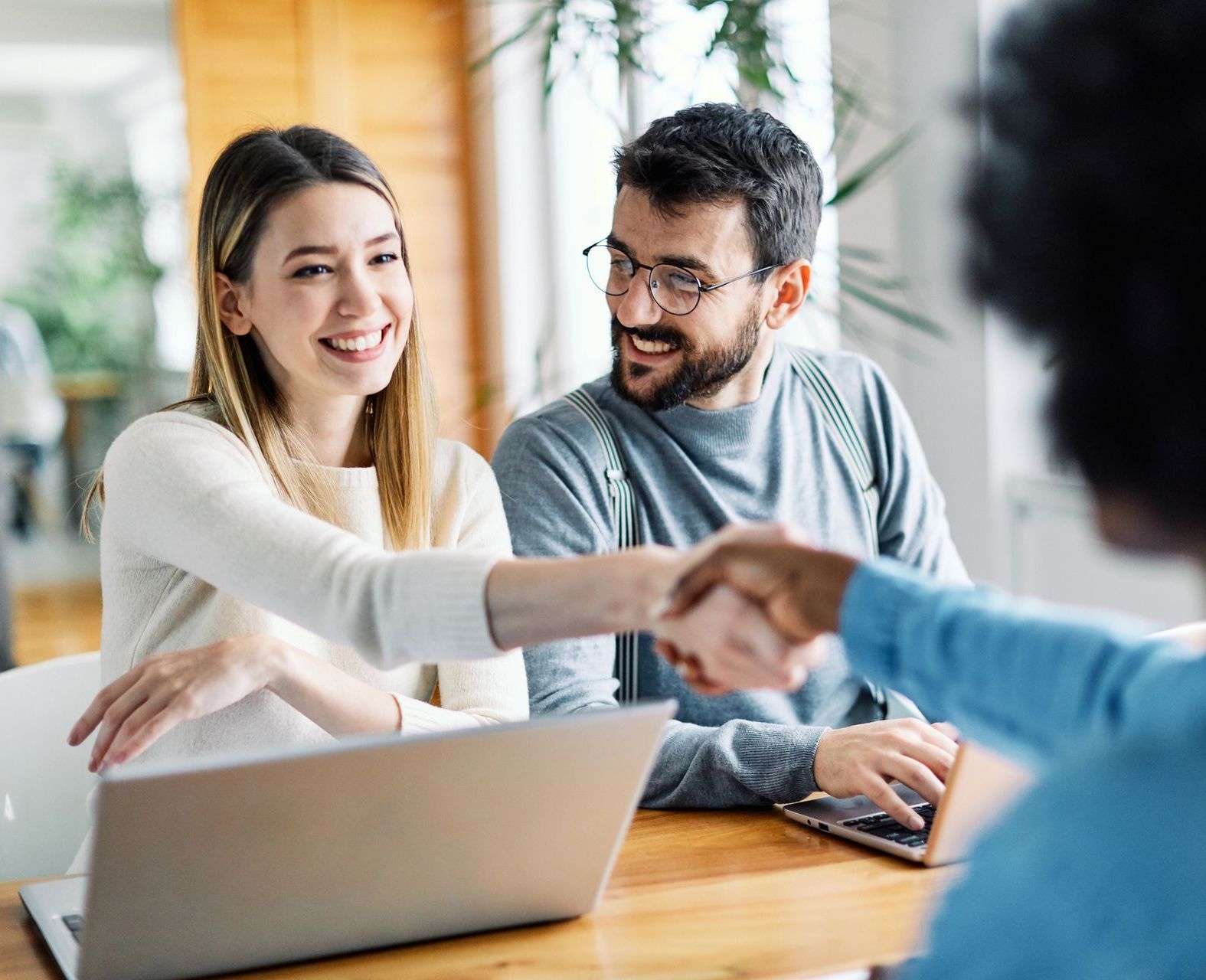 A man and a woman are shaking hands while sitting at a table with a laptop.