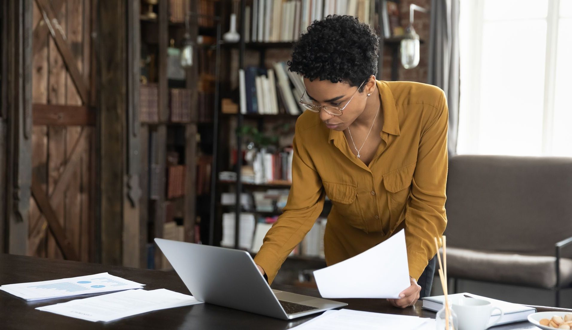A woman is standing at a desk looking at a laptop computer.