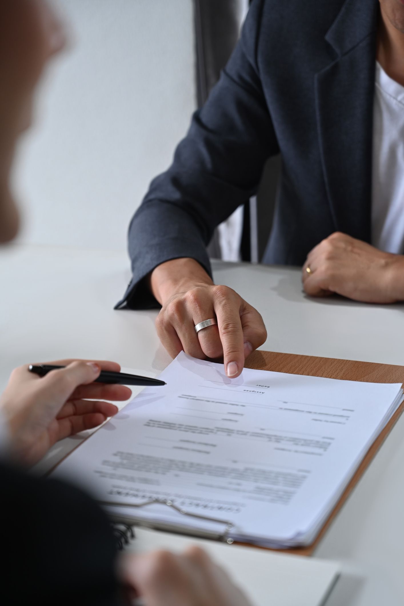 Person pointing at document on clipboard, another holding pen, at a table.