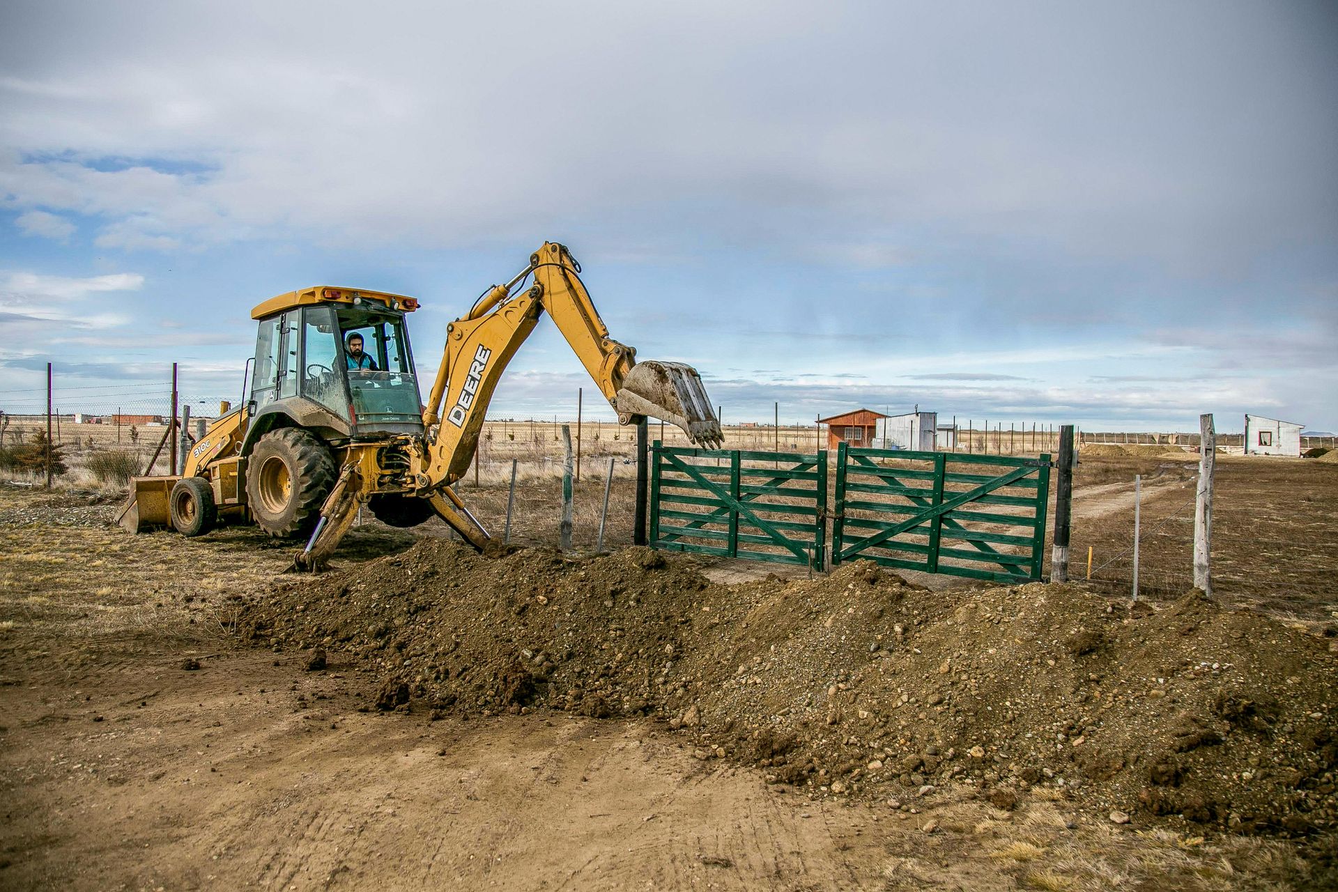 Backhoe loader digging soil beside a green farm gate in a fenced field