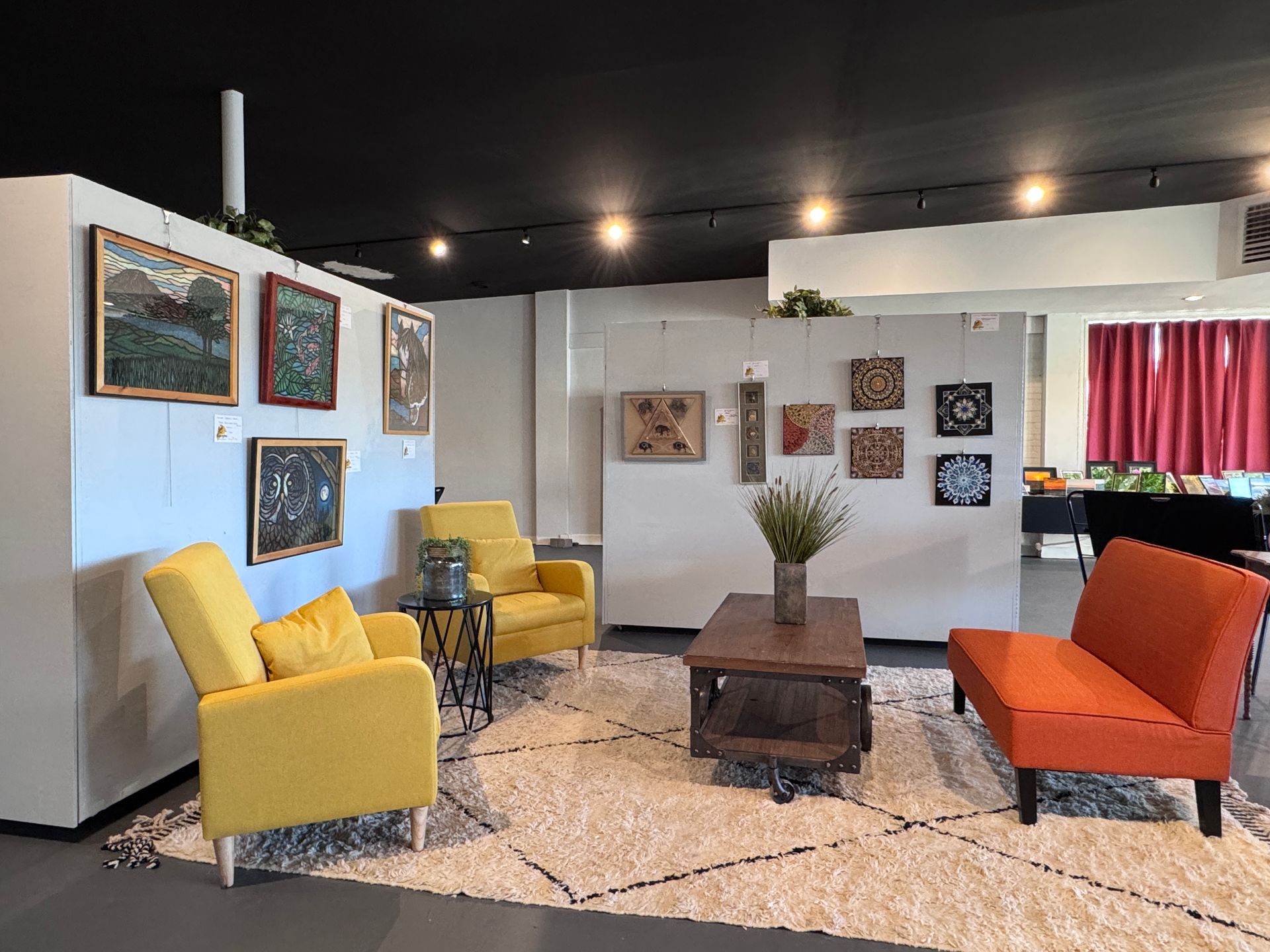 Art gallery interior with framed artwork, seating areas (yellow and orange), and a white rug.