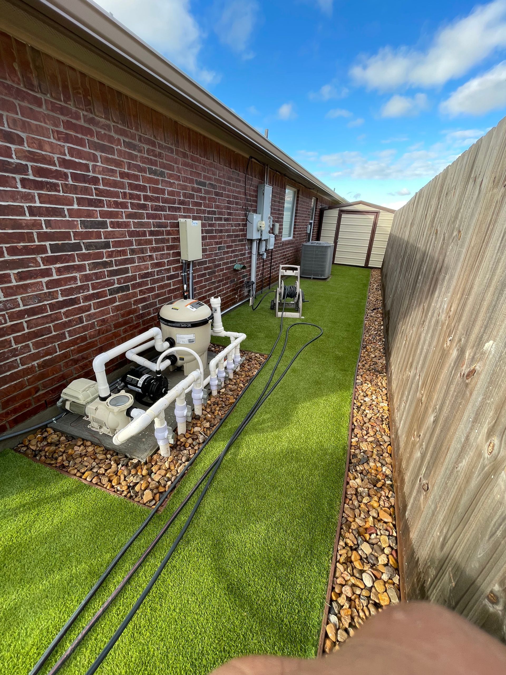 Backyard utility units beside brick house and fence, with gravel border and artificial grass strip