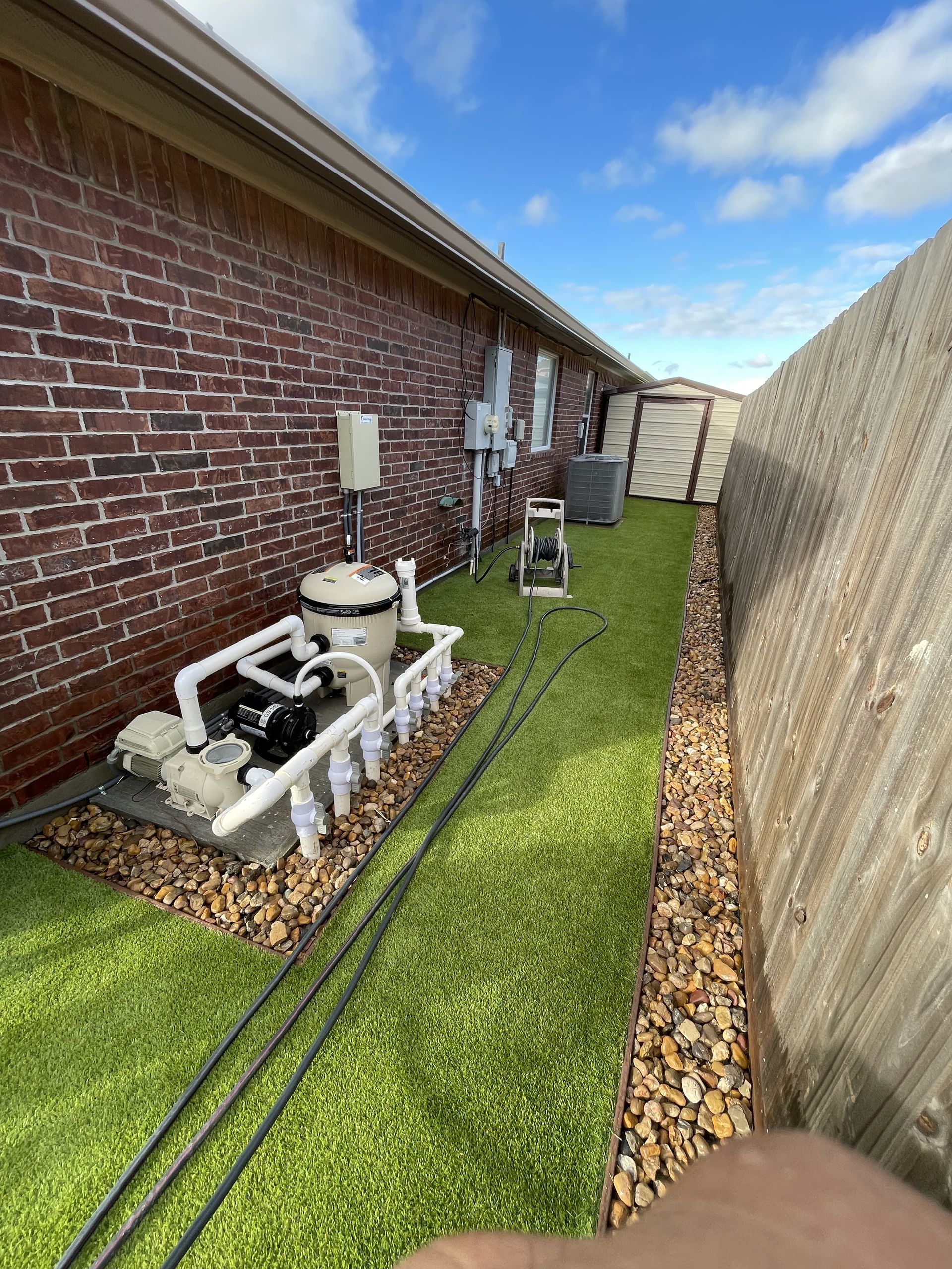 Pool pump equipment sits on gravel next to a brick house wall, with an artificial turf yard and wood fence alongside.