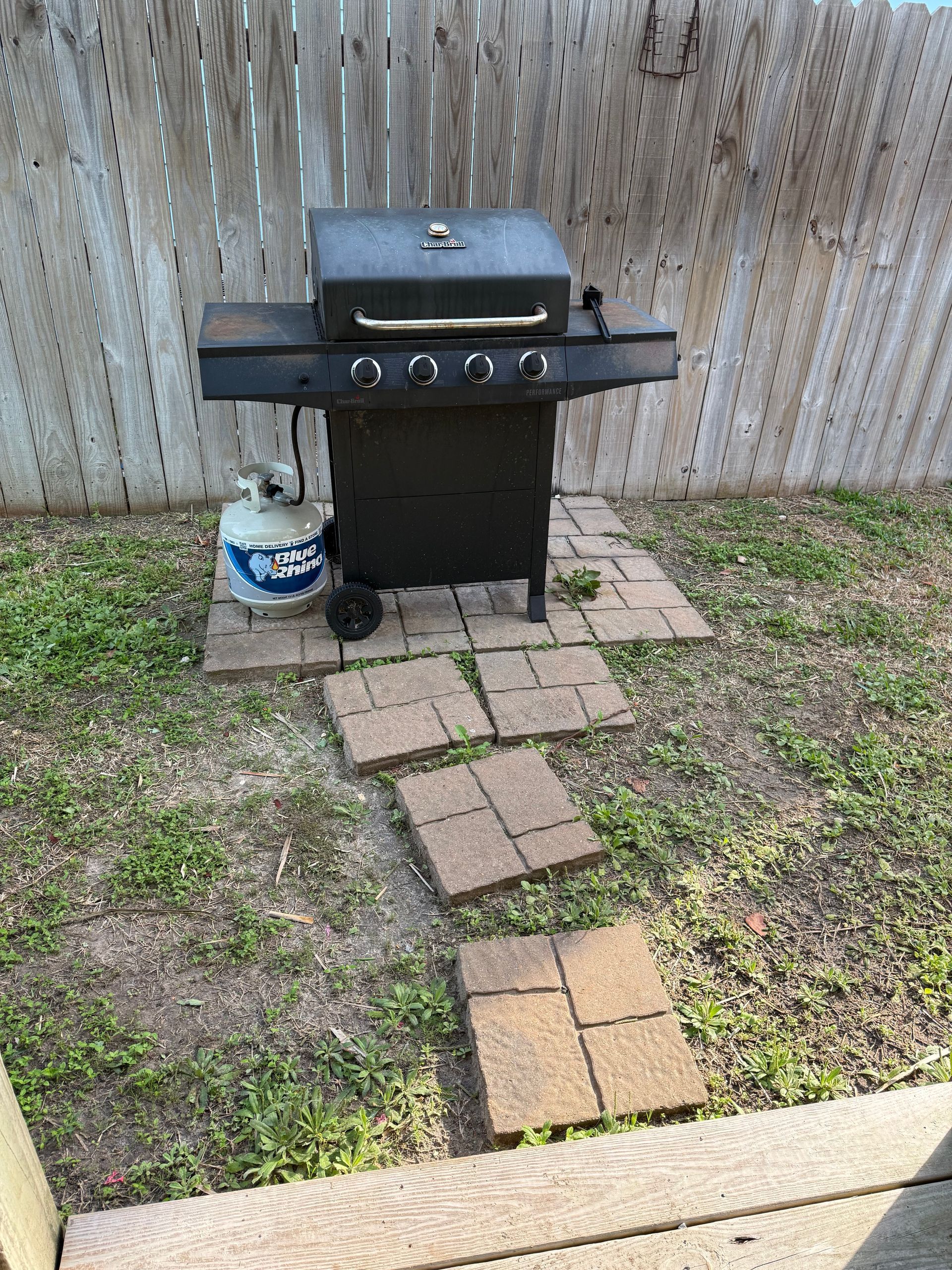 A black propane grill sits on a brick patio in a grassy backyard against a wood fence.