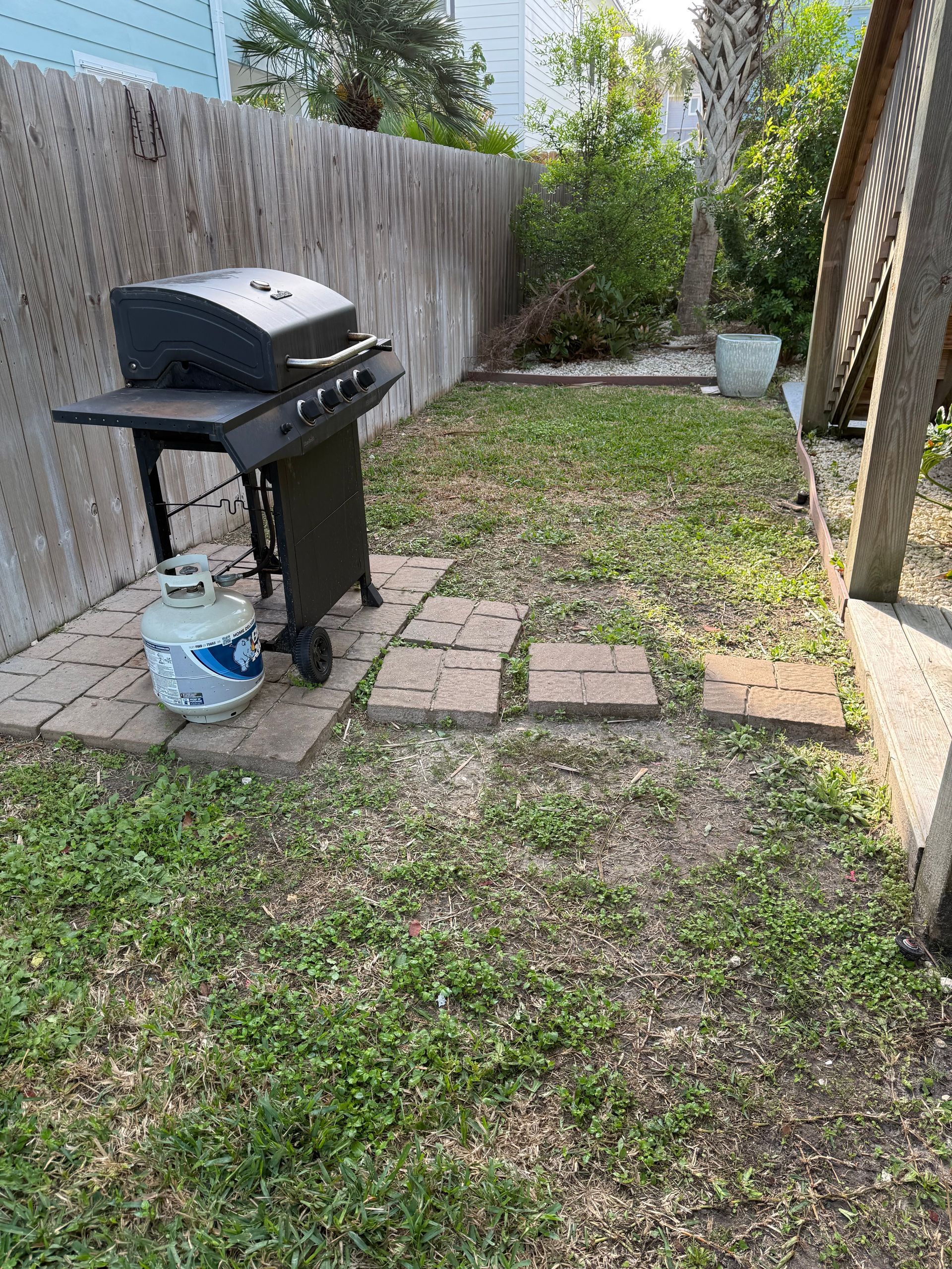 A black gas grill with a propane tank sits on a brick patio in a backyard with patchy grass near a wooden fence.