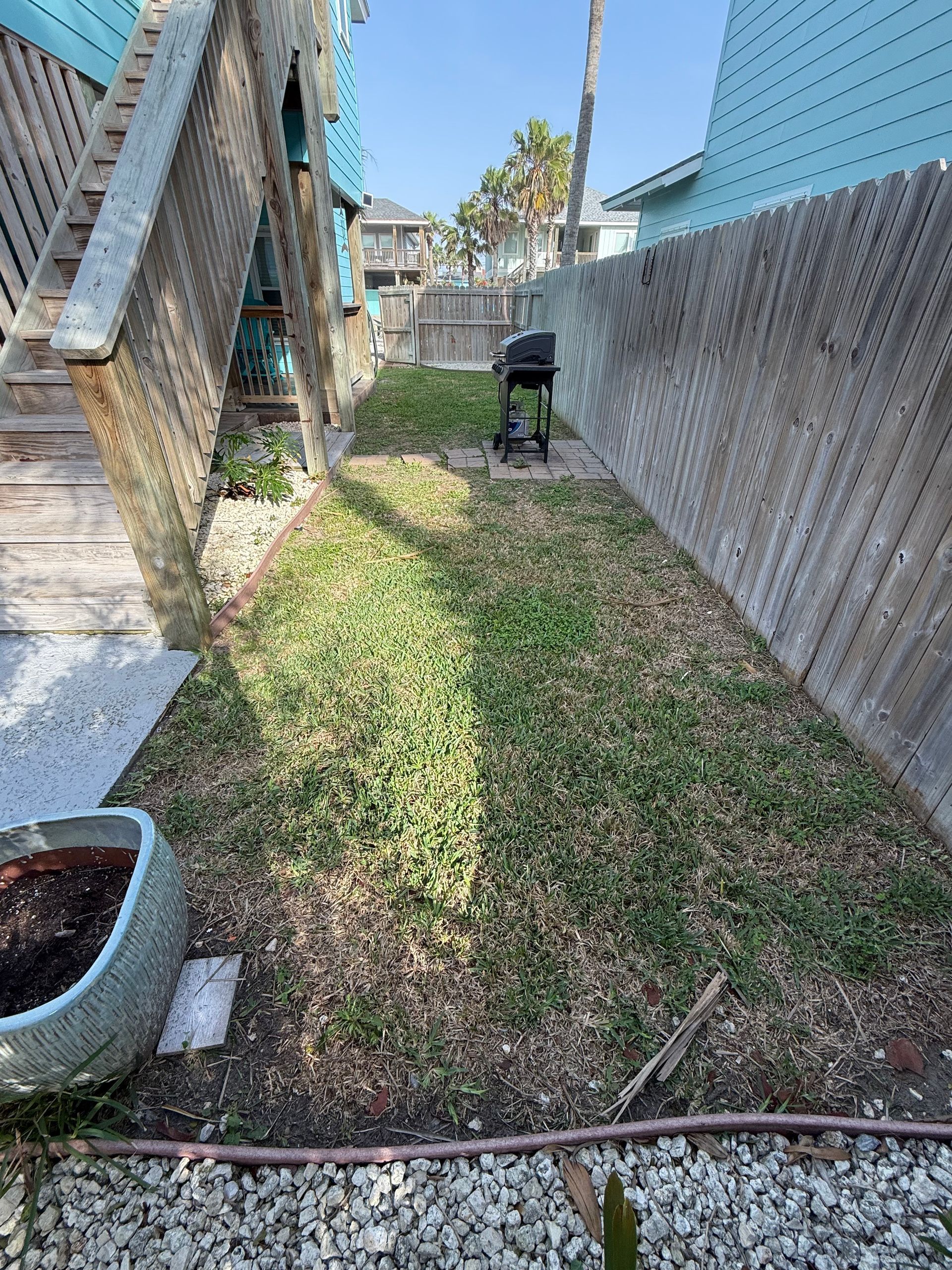 A narrow, sunny backyard between a wooden house staircase and a tall wooden fence, with a grill at the far end.