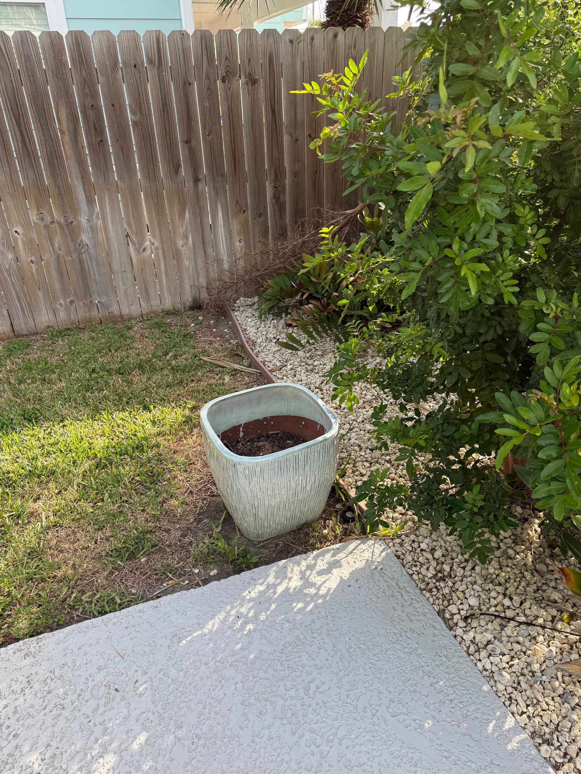 A light gray square planter sits on a bed of gravel near a wooden fence and a leafy green bush in a backyard.