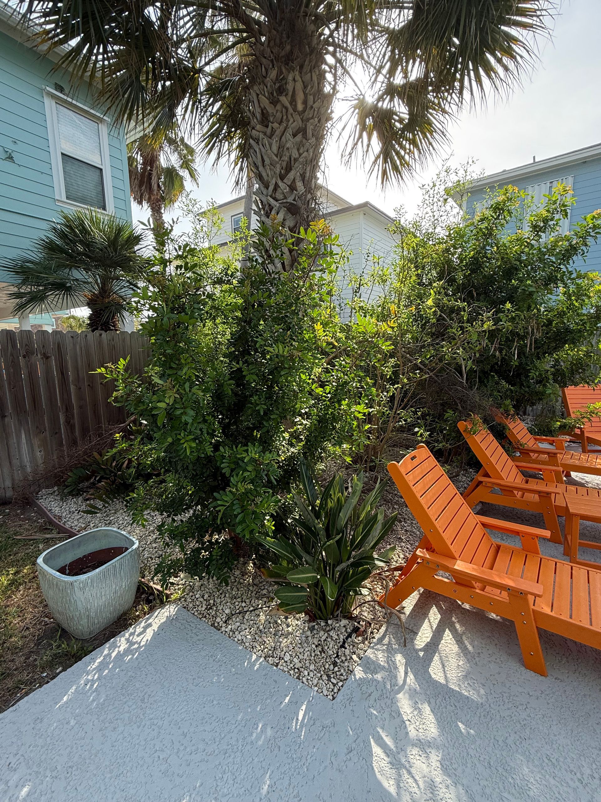 A sunlit patio with orange lounge chairs, lush greenery, and a palm tree against light blue beach houses.