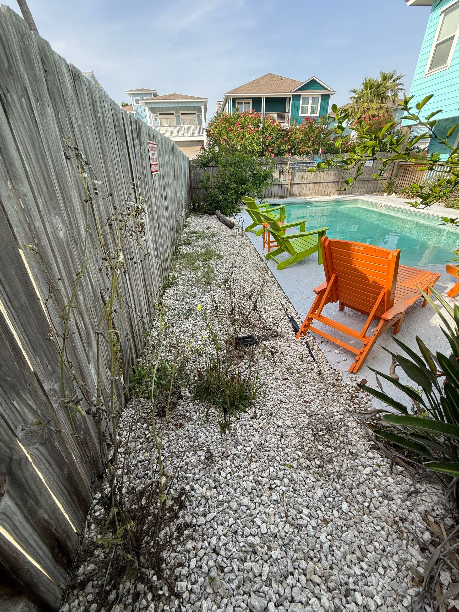 A small backyard with a pool, two Adirondack chairs (one lime green, one orange), and a gravel area beside a wood fence.