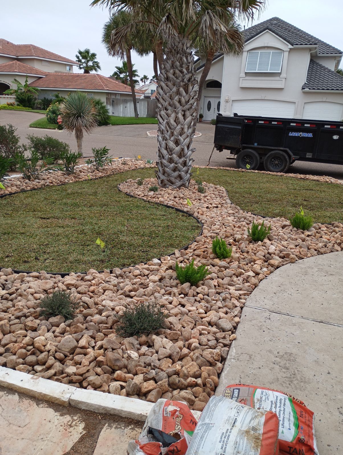 A landscaped front yard with tan rocks, small shrubs, a palm tree, a patch of grass, and a dumpster in the driveway.