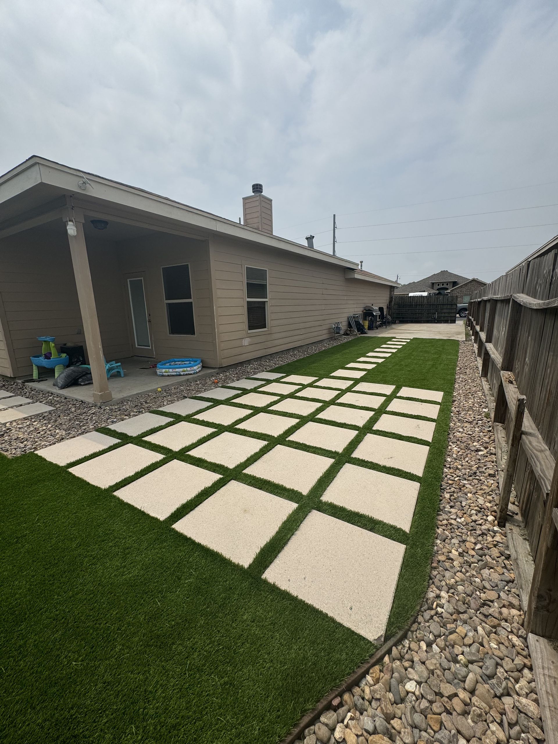 A backyard featuring a square-tiled path set in synthetic turf, bordered by rocks next to a tan house with a covered patio.