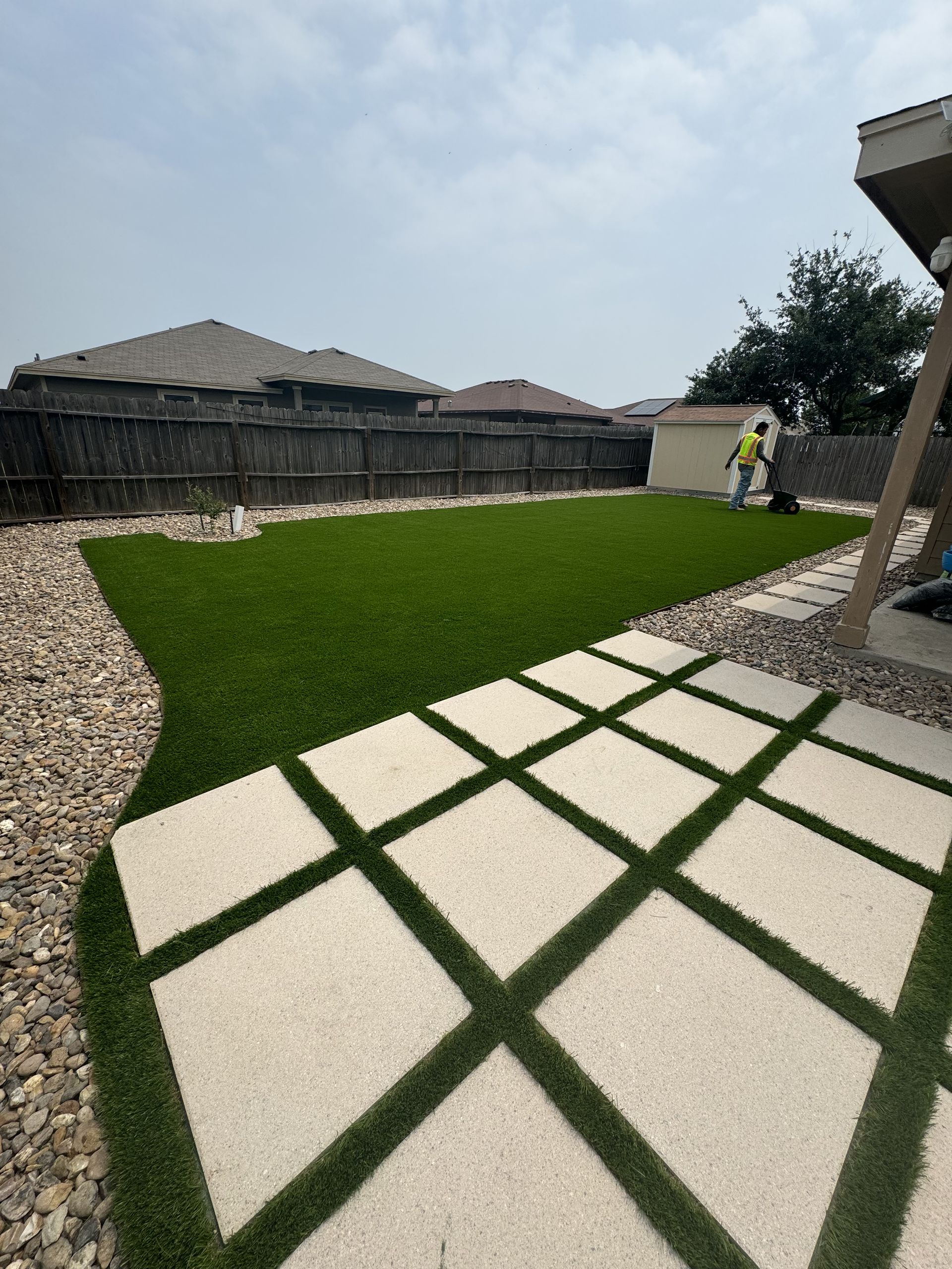 A backyard featuring a square-tiled patio with turf inlays, a large green artificial lawn, and gravel borders.