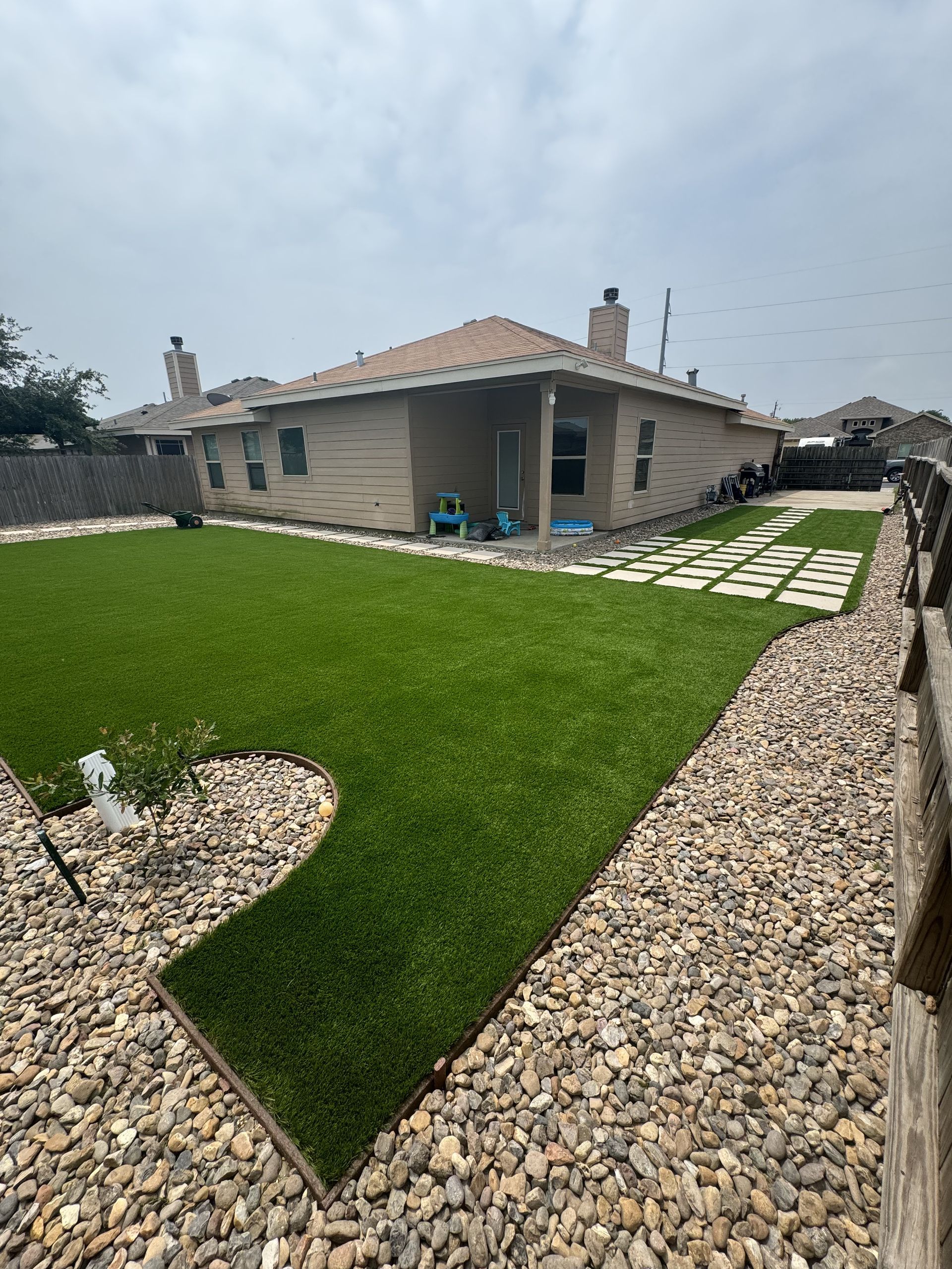 A backyard with green artificial turf, surrounded by light-colored landscaping rocks and a path of square pavers.