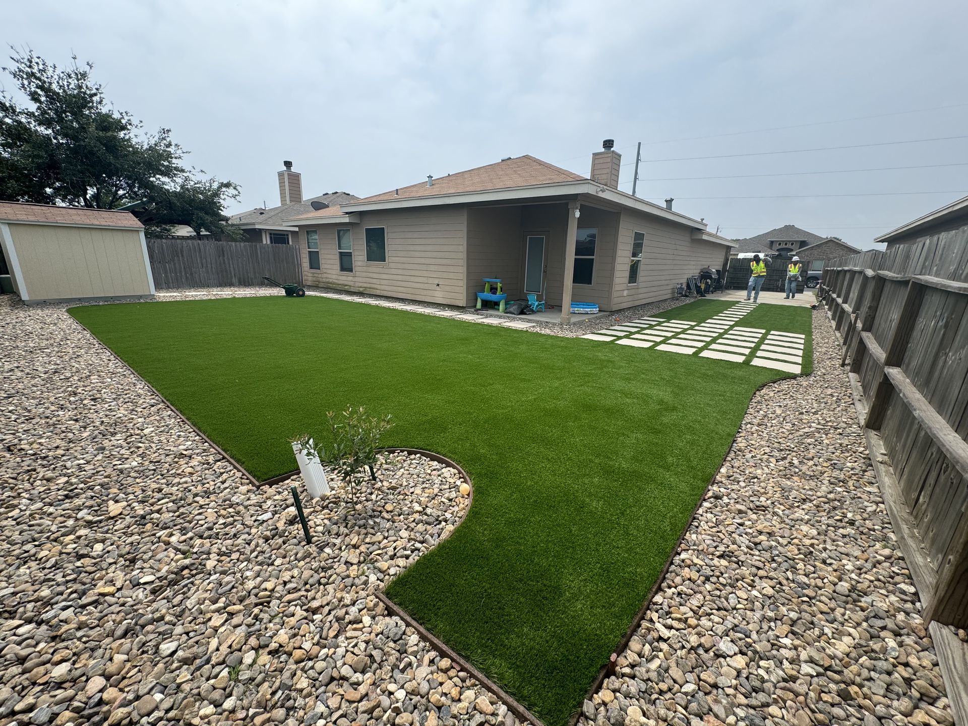 A tan house with a rectangular patch of bright green artificial turf surrounded by landscaping rocks in a backyard.