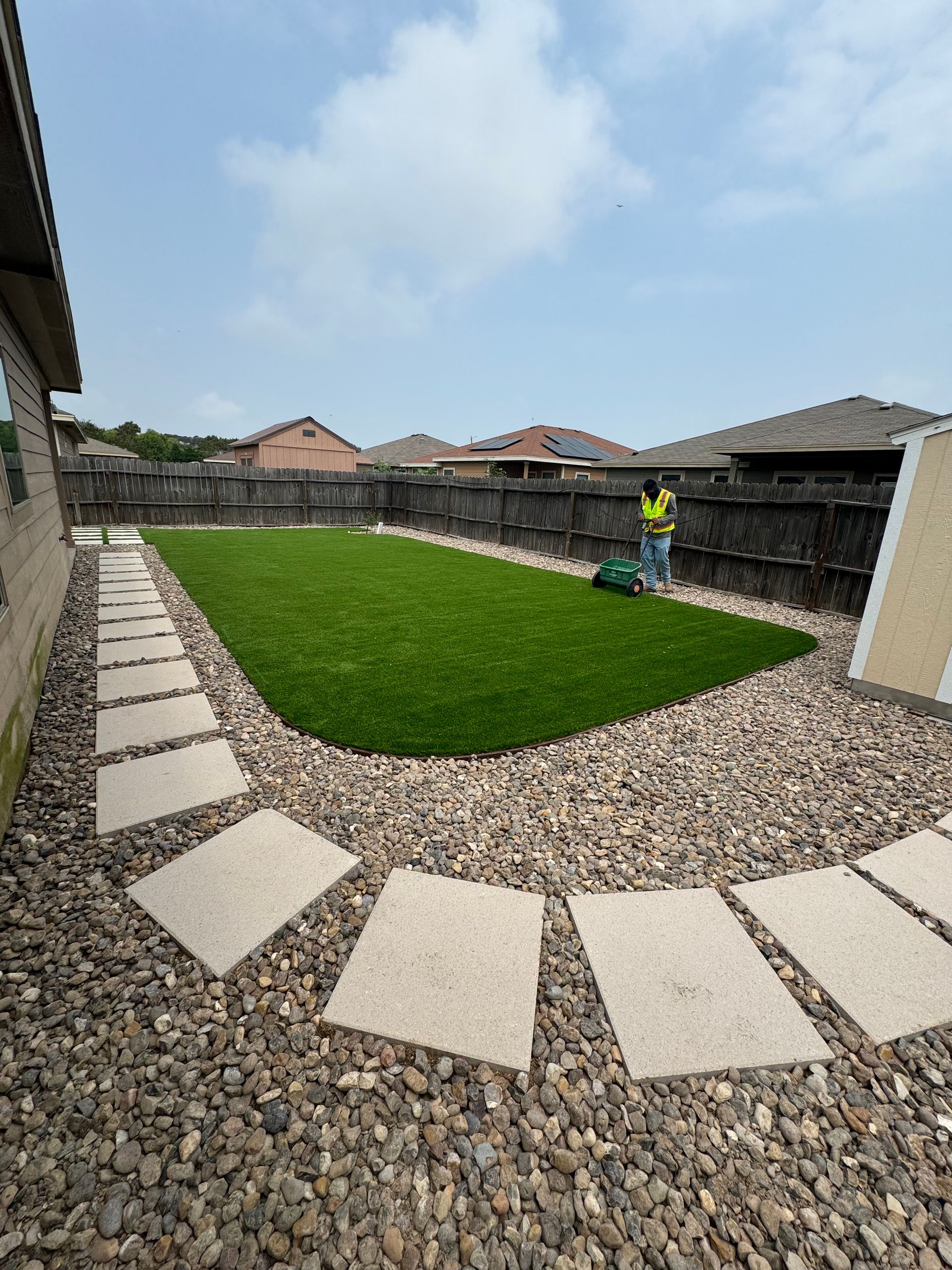 Curved gravel yard with stepping stones, green lawn, and a worker in a bright yellow shirt near a fence.