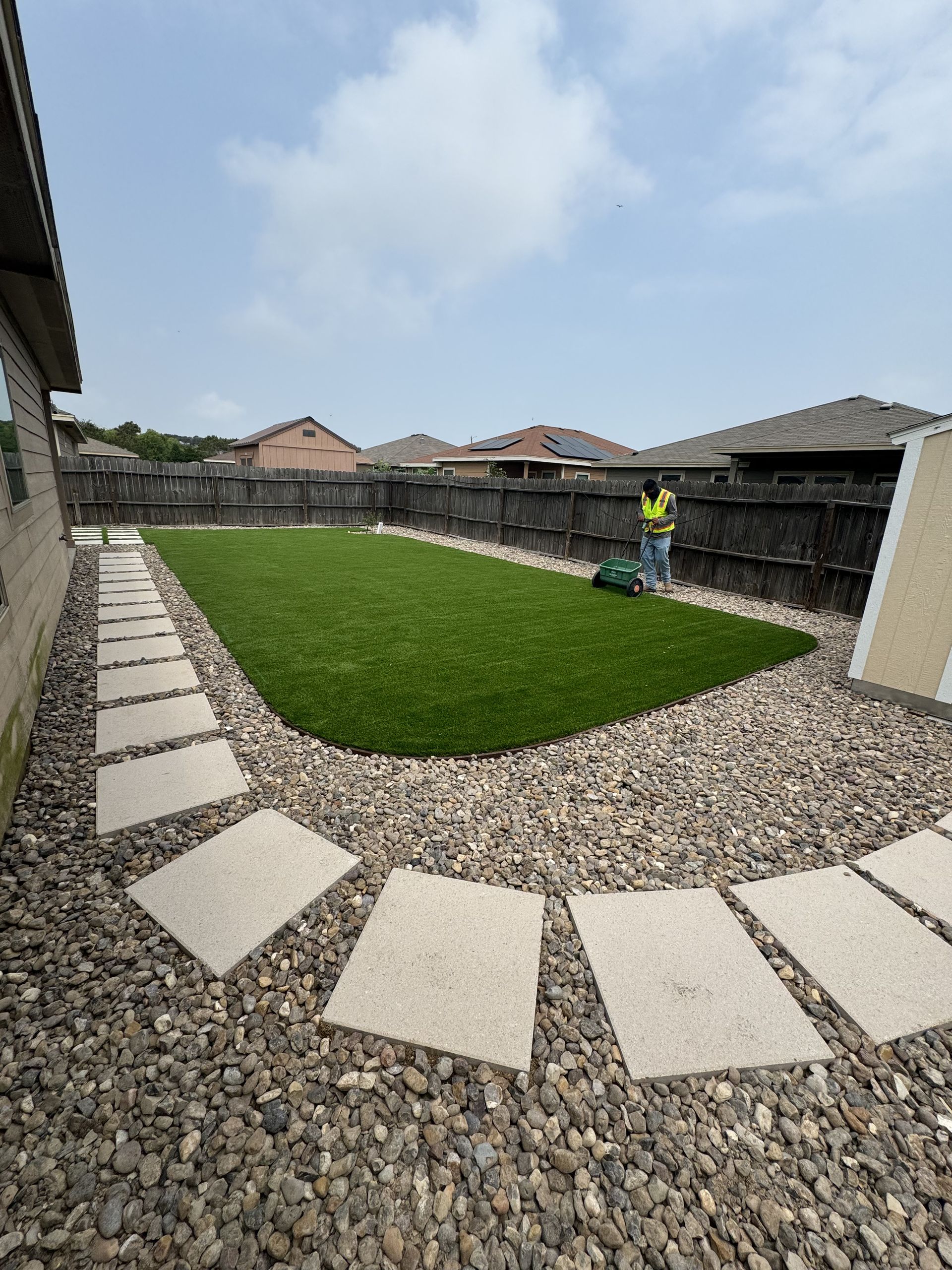 A worker in a high-visibility vest stands by a small patch of artificial grass surrounded by stone pavers and gravel.