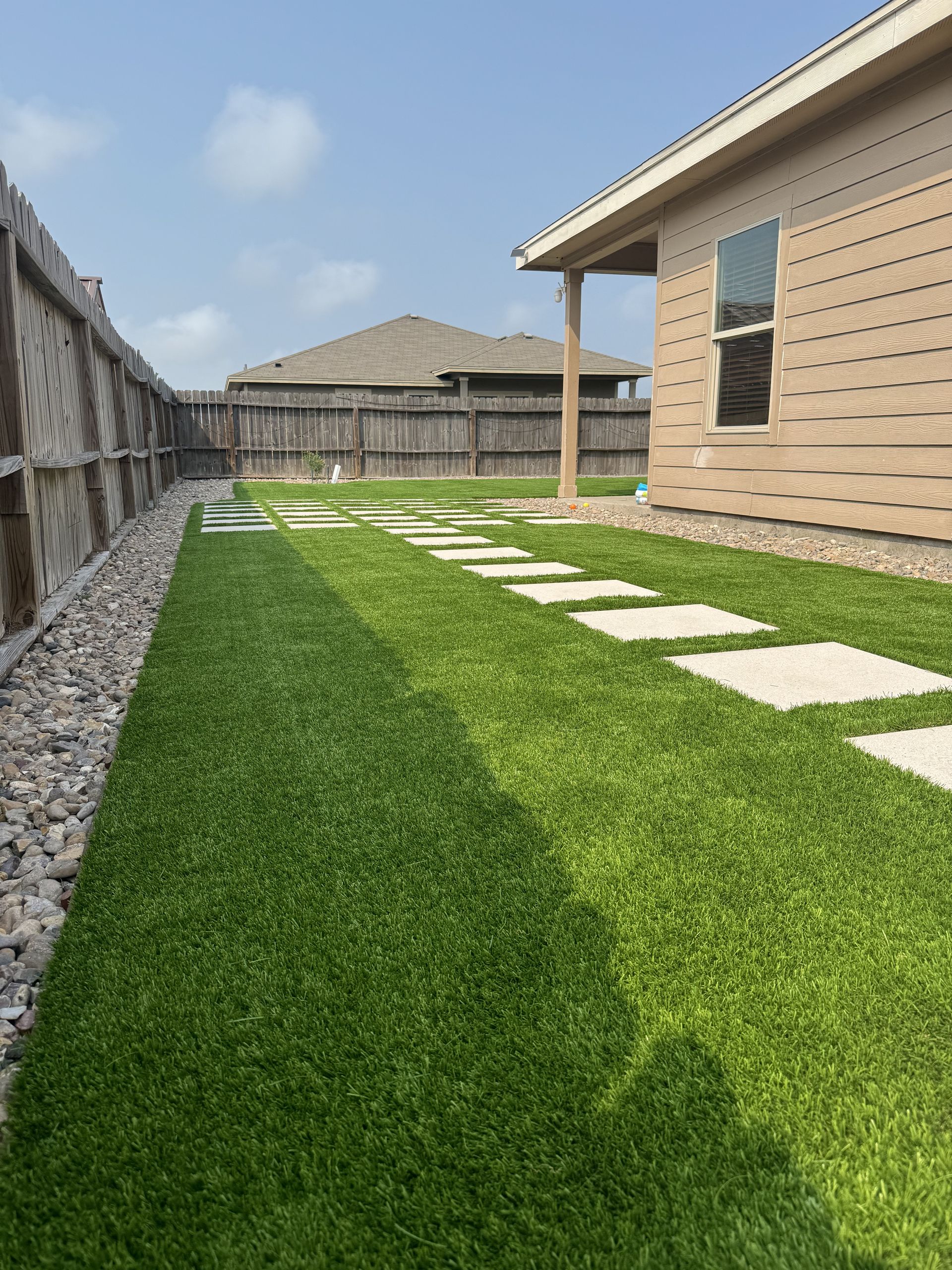 A backyard with green artificial turf, a rock border along a wooden fence, and a stone path leading to a tan house.