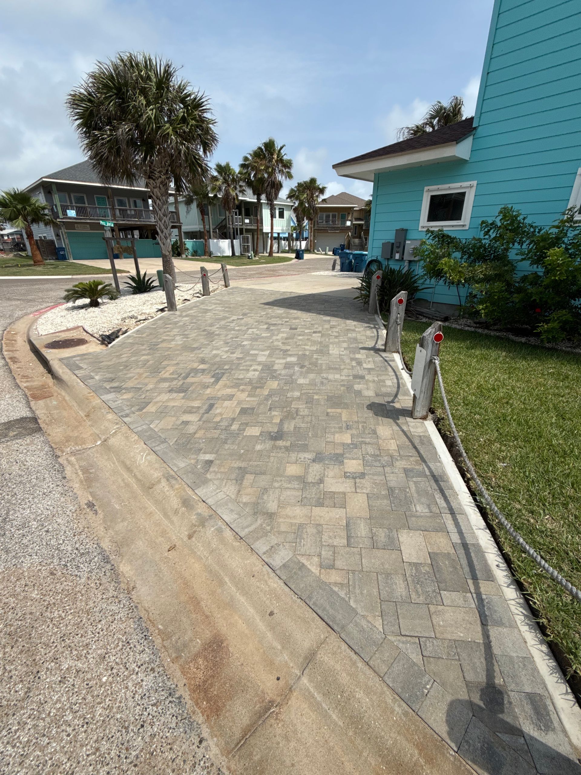 A paved brick driveway leading toward a blue house, lined with posts and rope, under a clear sky.