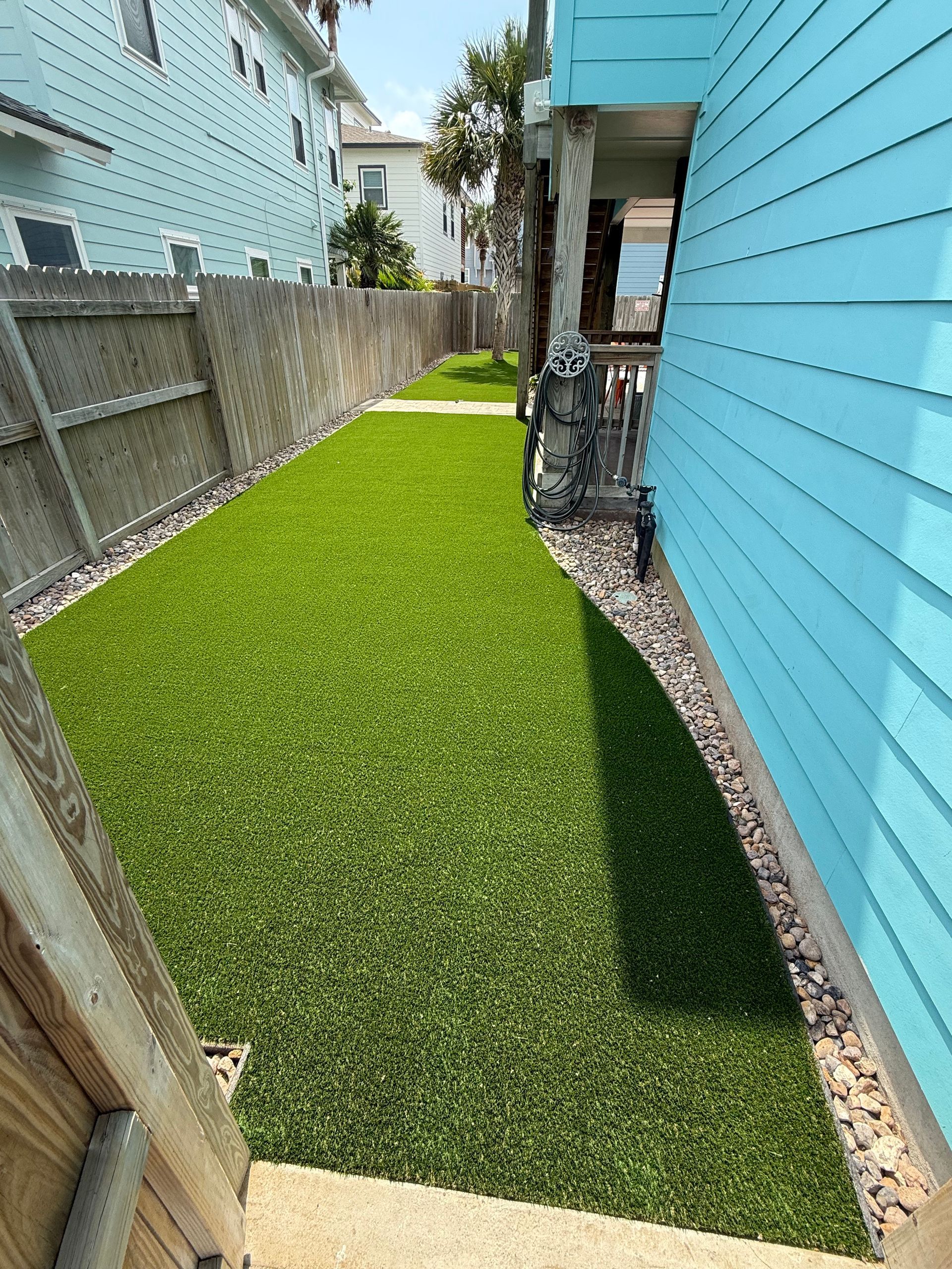 A narrow yard with bright green artificial turf between a turquoise house and a wooden fence, with gravel border.