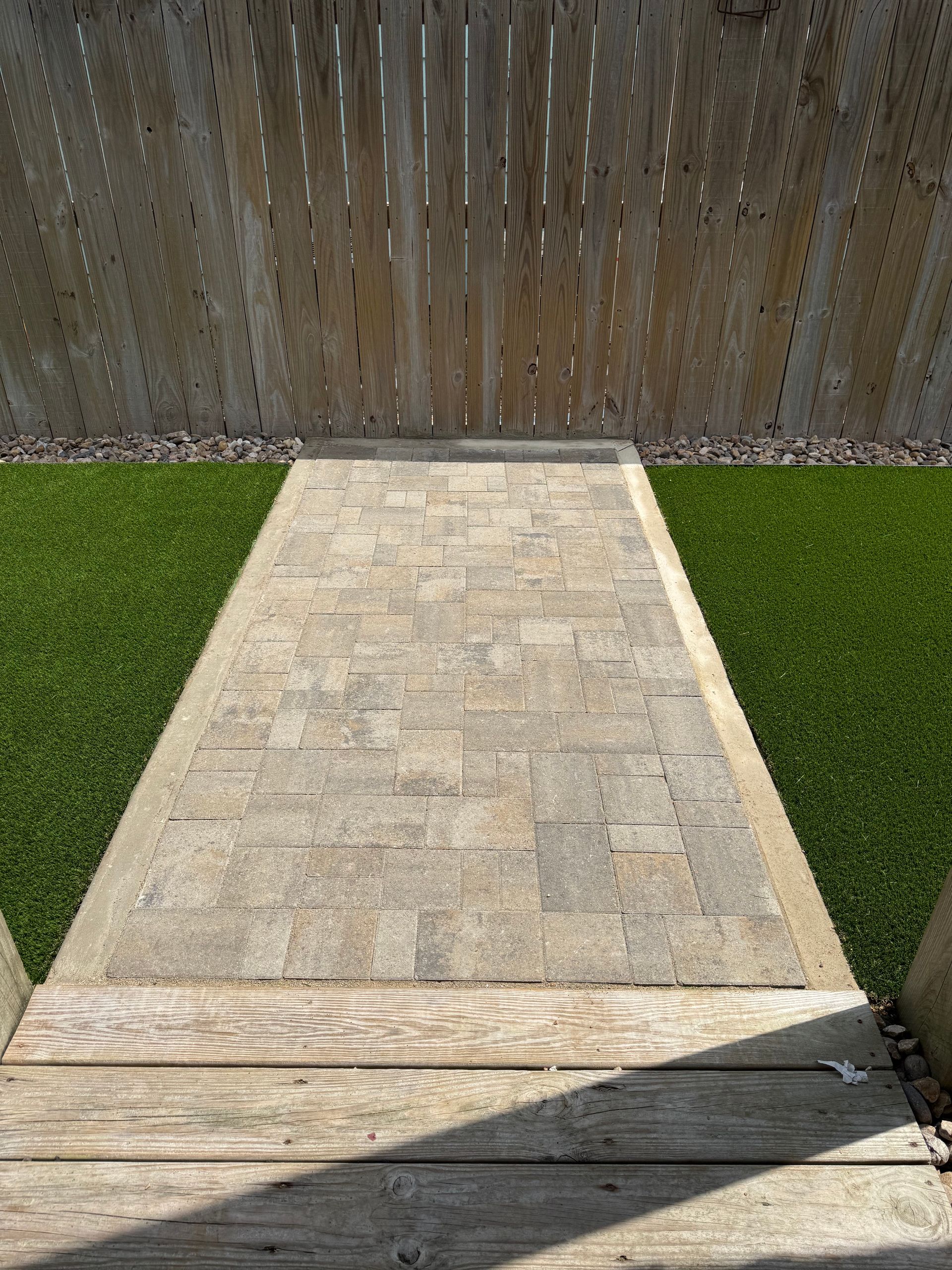 A paved walkway made of rectangular stone tiles leads from a wooden deck toward a wooden fence in a yard with green grass.