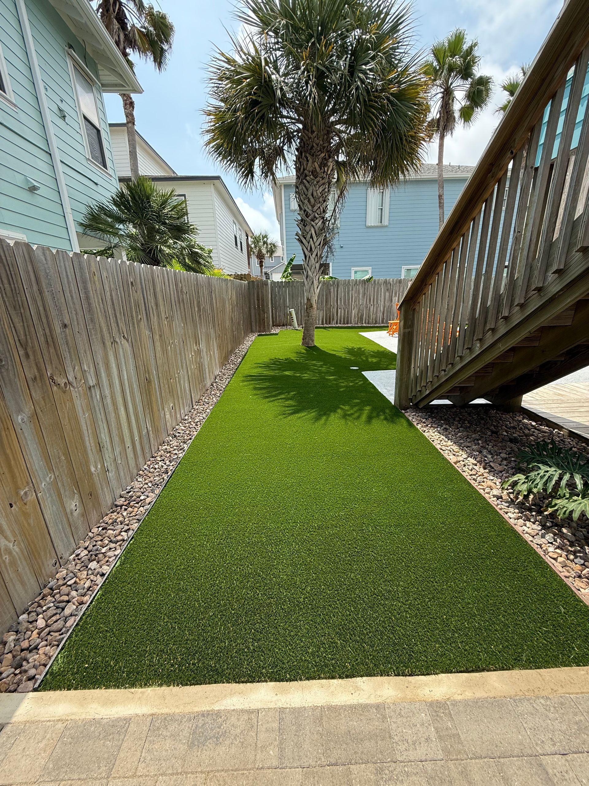 A narrow strip of artificial green grass between a wood fence and wooden stairs, with a palm tree in the background.