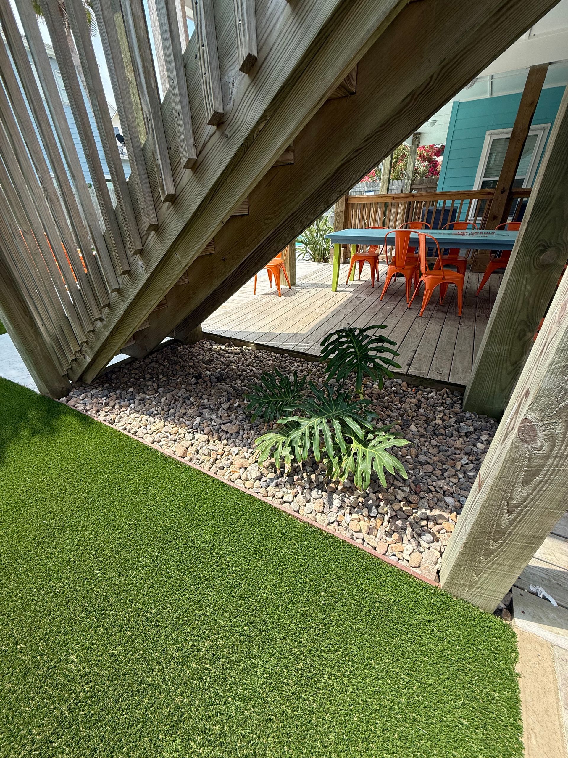 Stairs overlook a garden bed with a small plant in gravel, next to a patio with blue furniture on a sunny day.