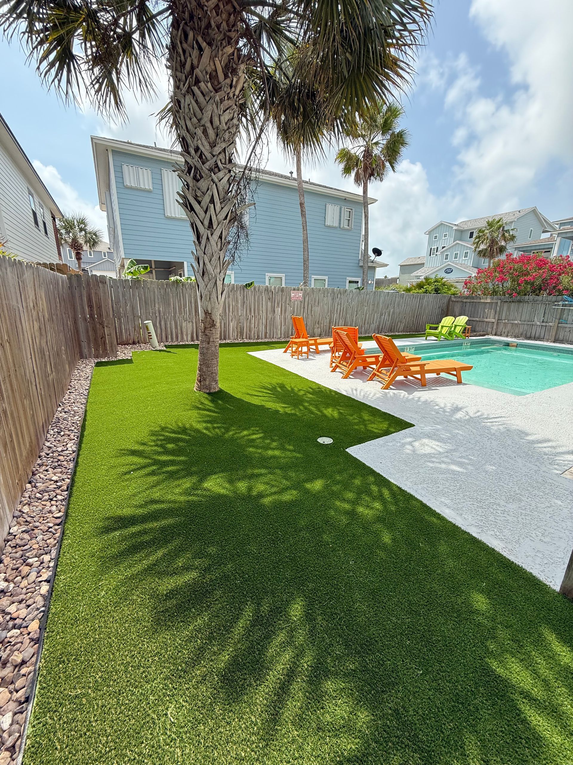 A backyard with synthetic green grass, a pool with turquoise water, and orange lounge chairs under palm trees.
