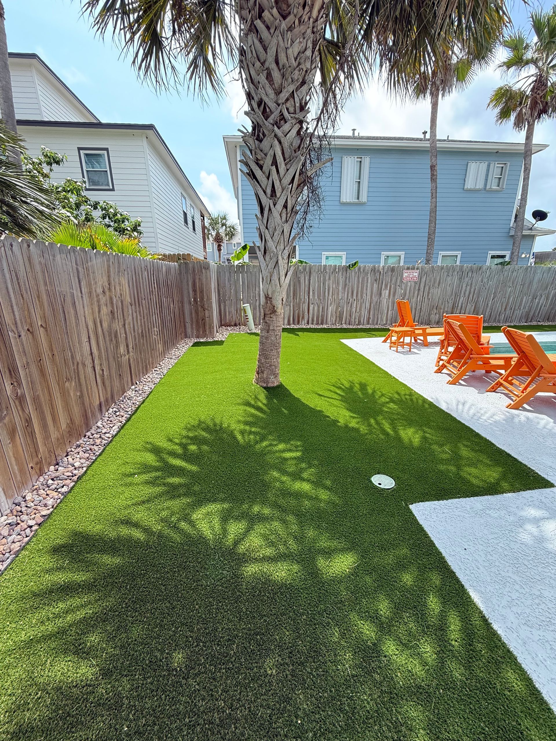 A backyard lawn with artificial grass, a central palm tree, and a patio with bright orange lounge chairs.
