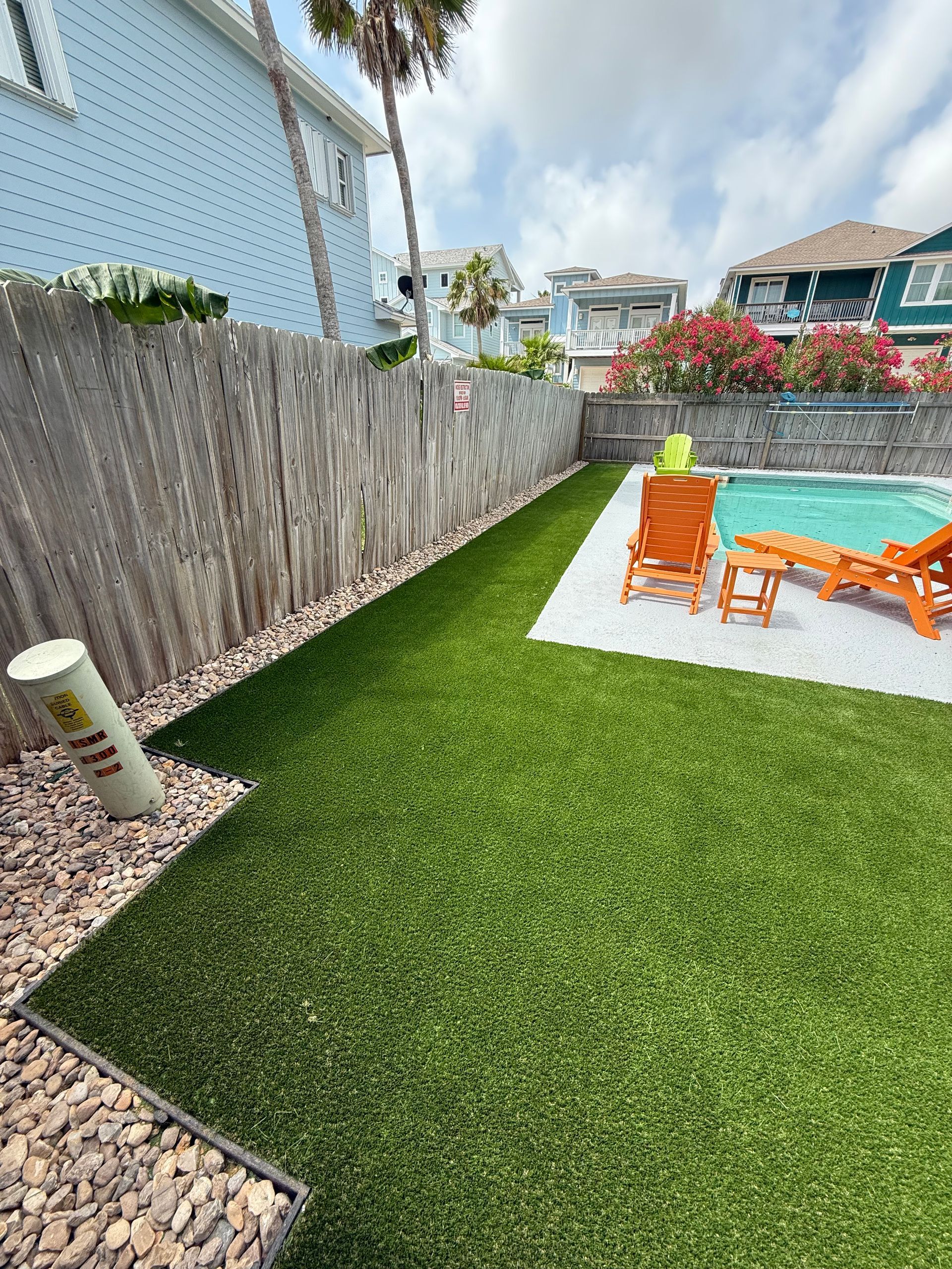 A backyard pool area with artificial grass, a wooden fence, and orange lounge chairs on a light-colored patio.