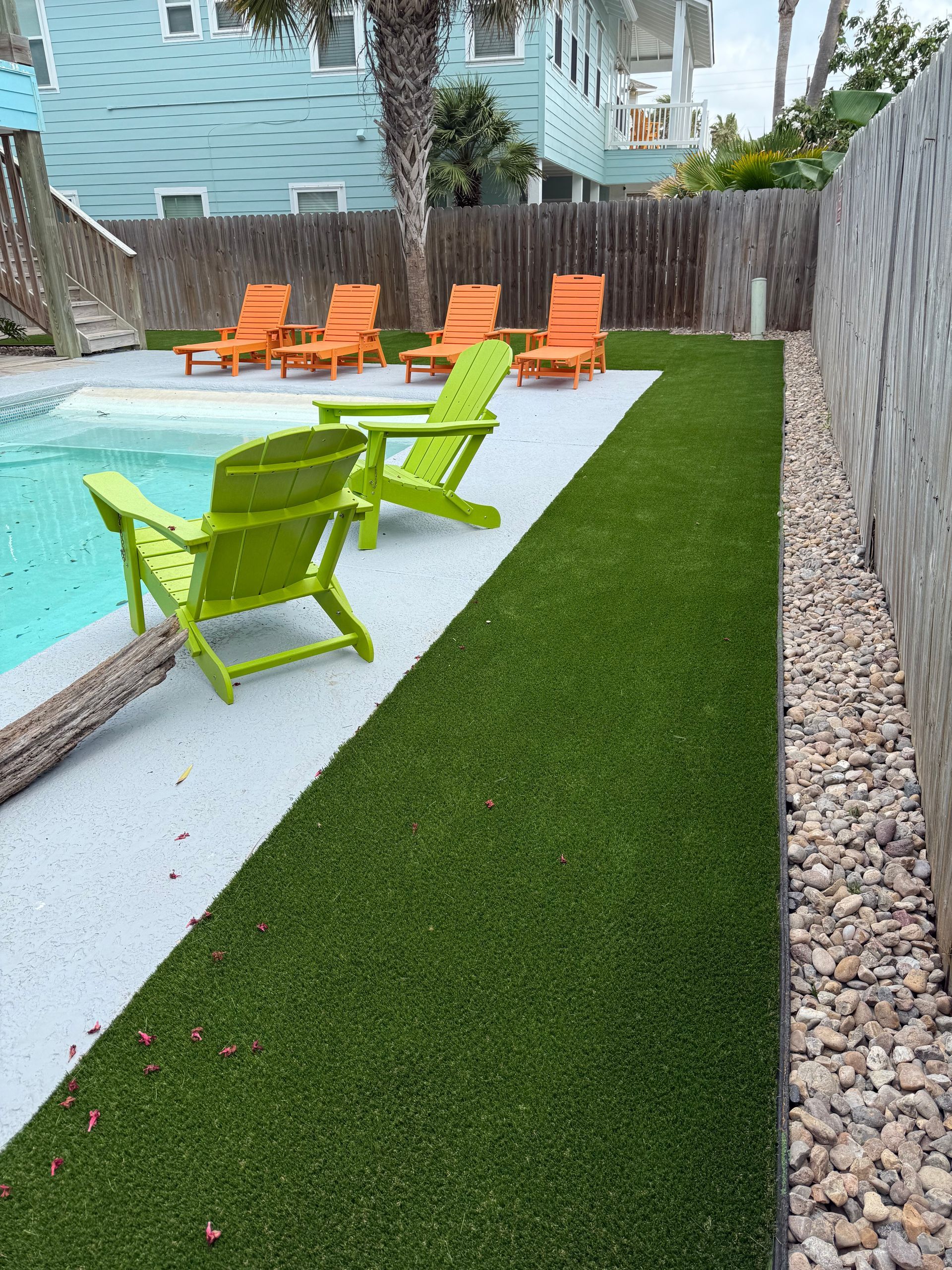 A swimming pool deck with two bright green Adirondack chairs, four orange lounge chairs, artificial turf, and gravel.