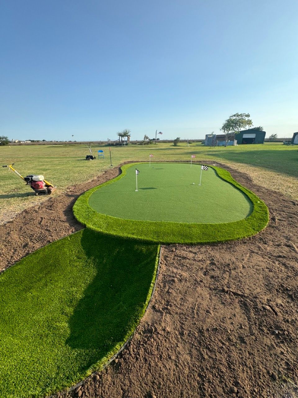 A putting green with artificial grass under a clear sky, surrounded by exposed dirt and a lawn mower to the left.