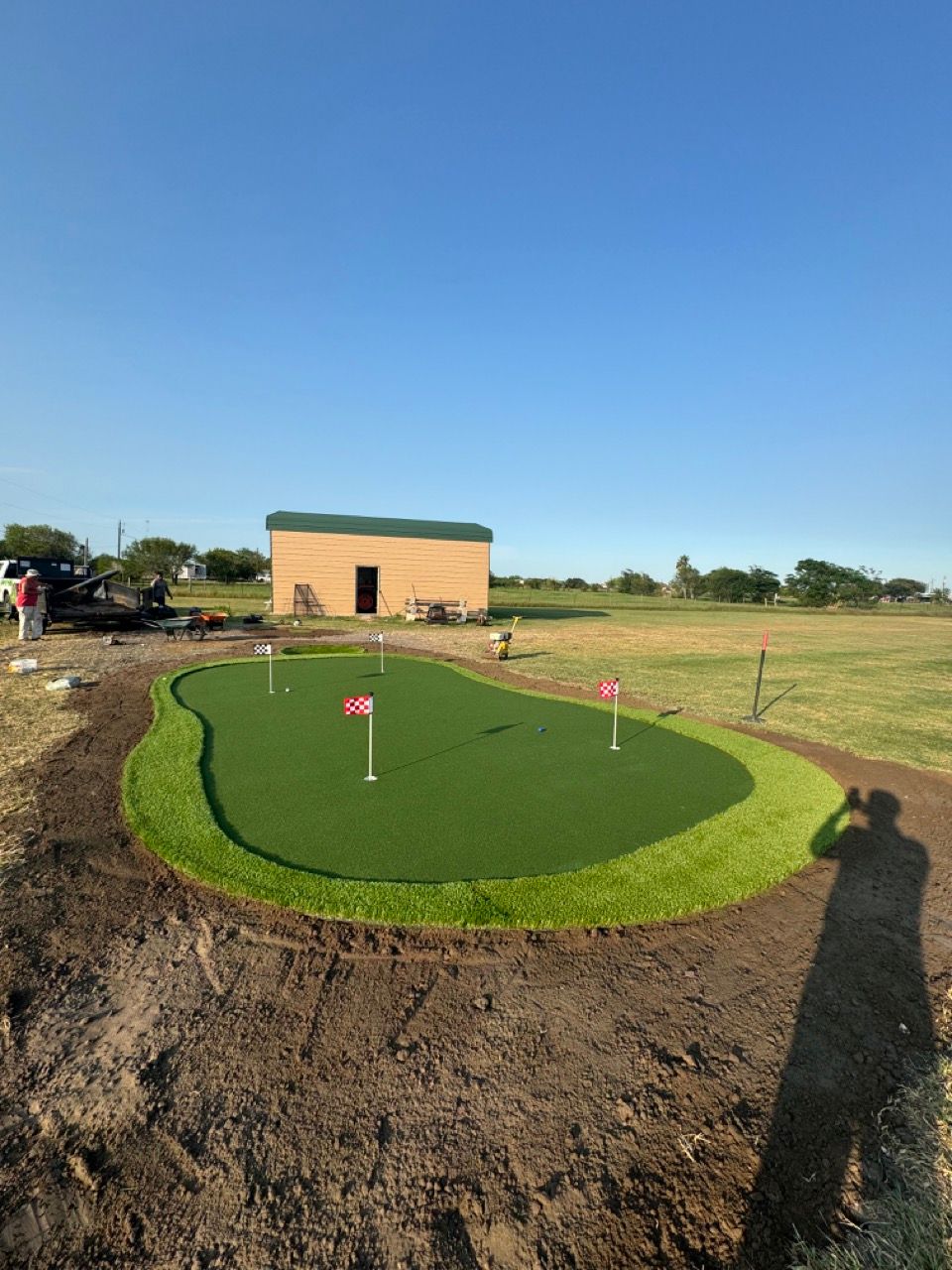 A backyard putting green with several holes and flags, surrounded by soil, in front of a small shed under a clear blue sky.