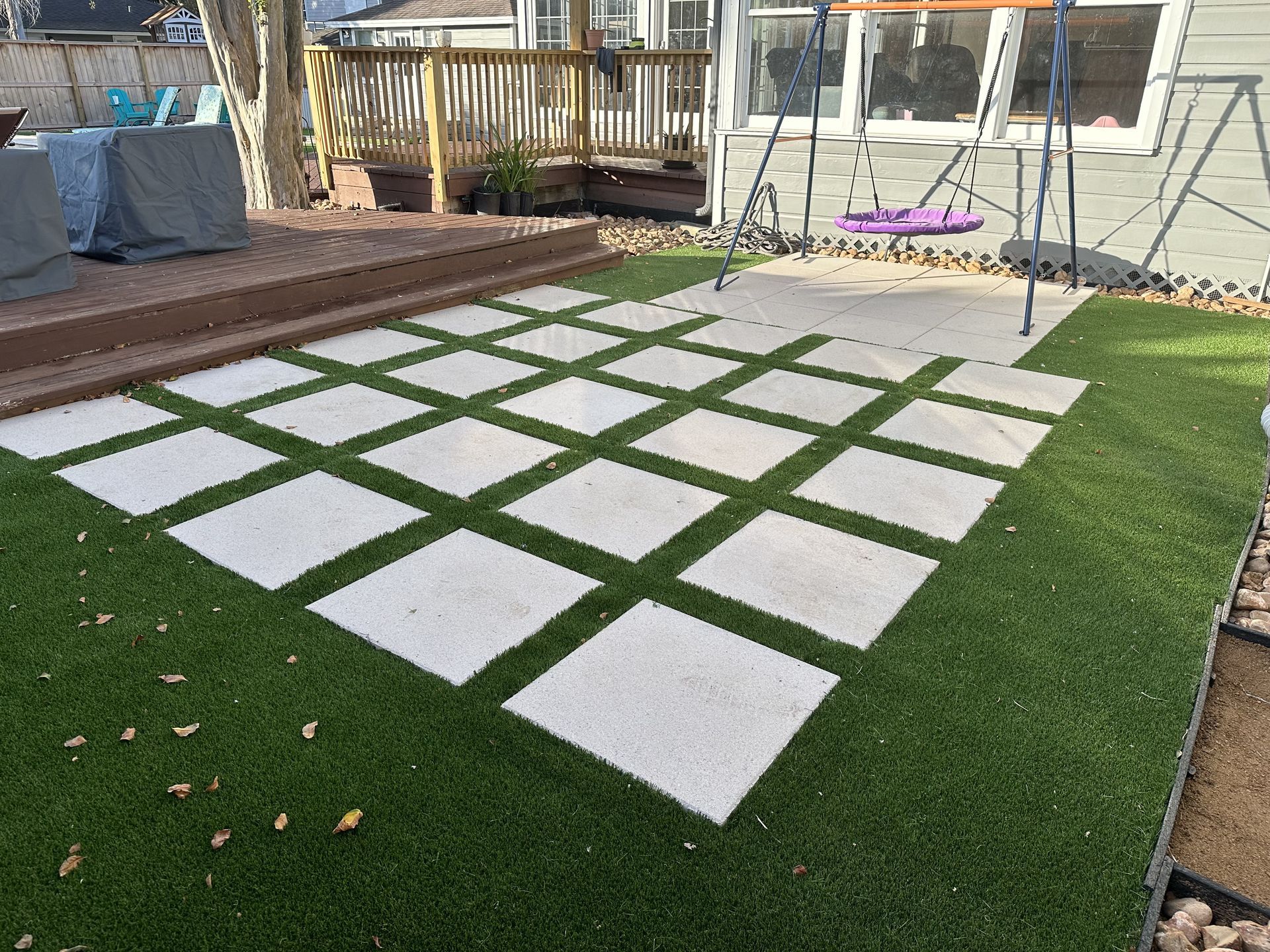 A square grid of white stone pavers set in artificial green turf, adjacent to a wooden deck and a swing set.