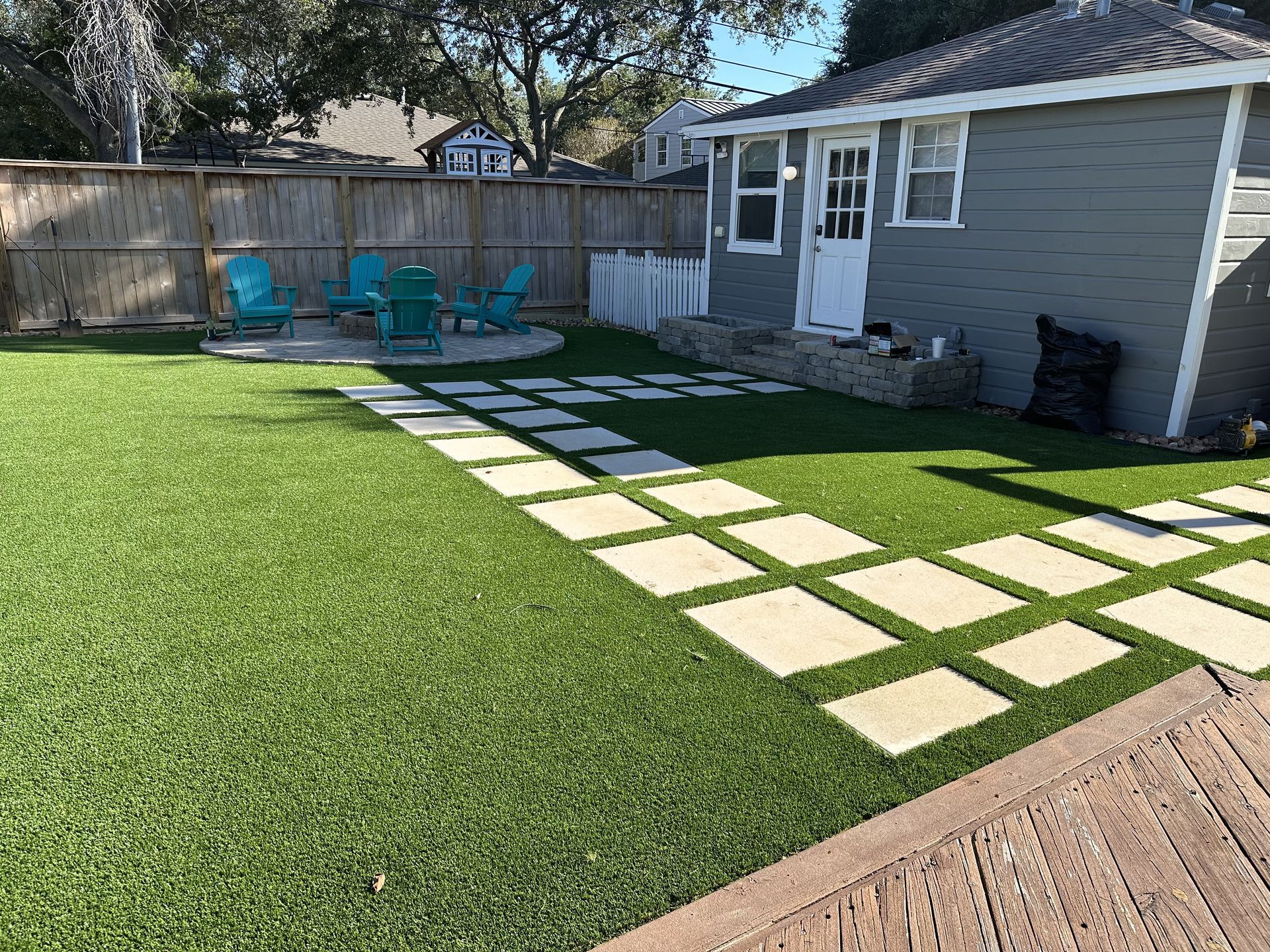 A backyard with green artificial turf, a paved stone walkway, and a small grey building near a wooden fence.