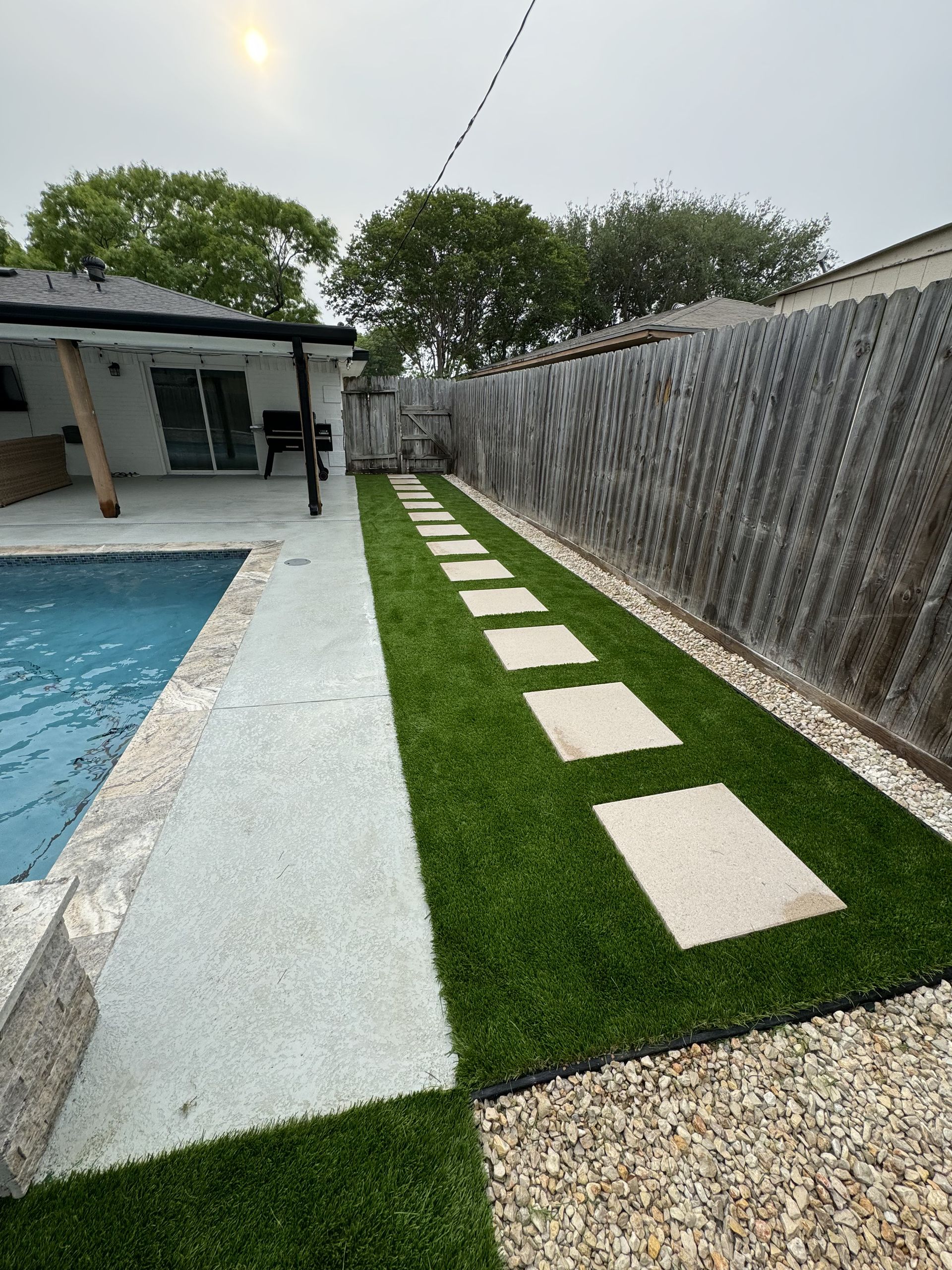 A backyard swimming pool patio featuring a grassy strip with square stone stepping stones next to a tall corrugated fence.
