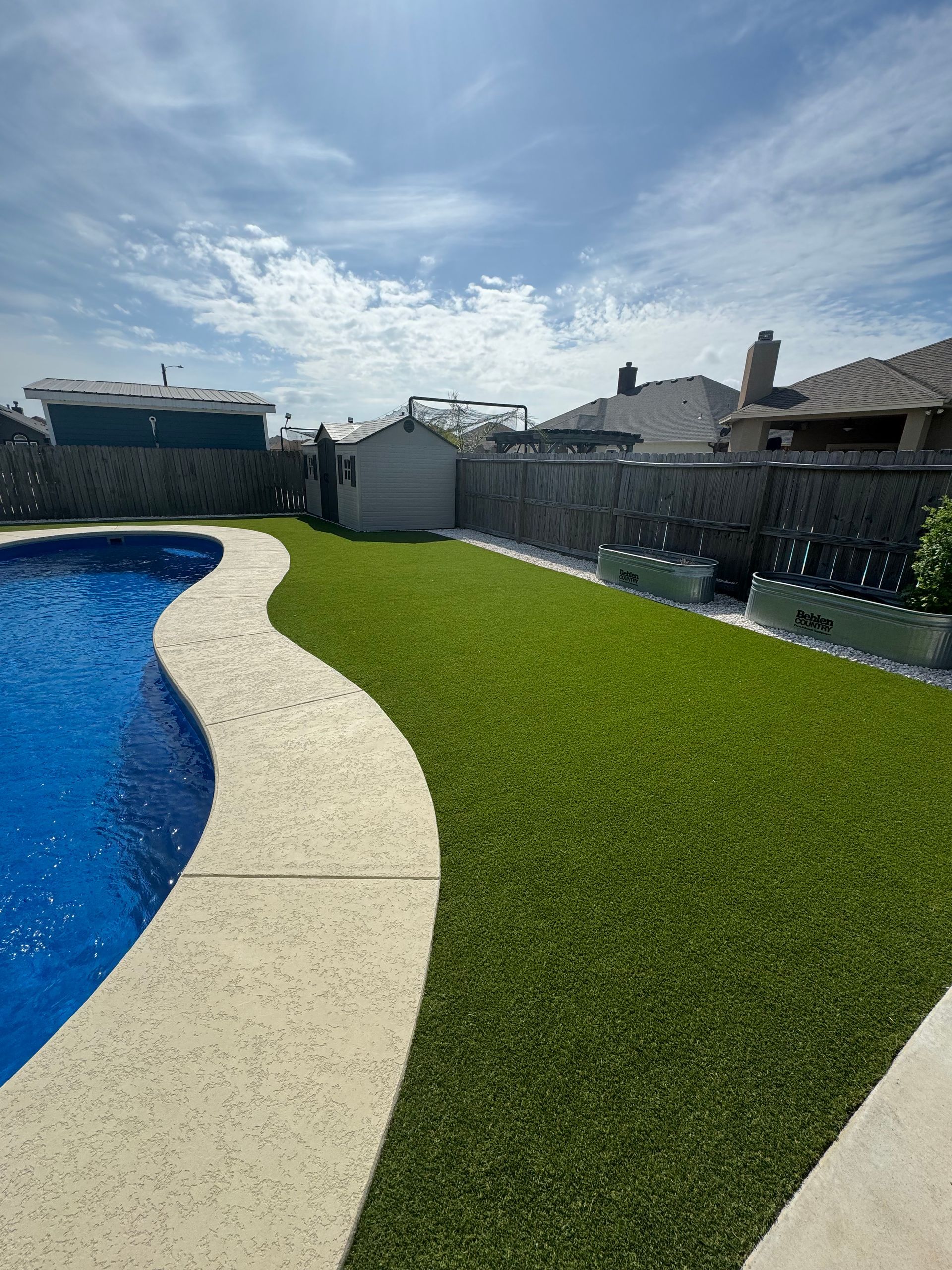 Backyard pool with curved white deck, green lawn, and fenced homes under a bright sky