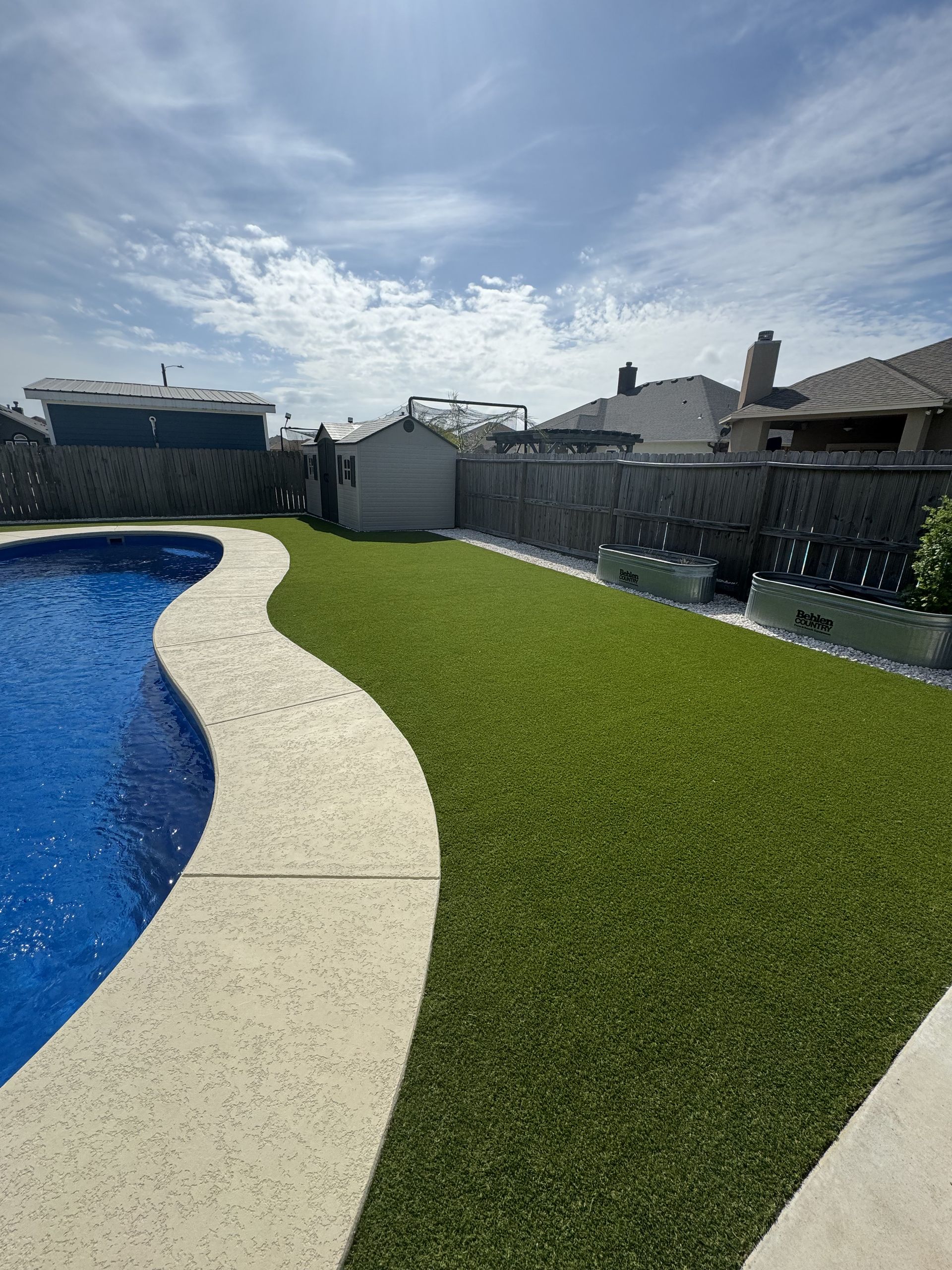 A bright blue swimming pool with a curved stone edge next to a patch of green artificial turf in a suburban backyard.