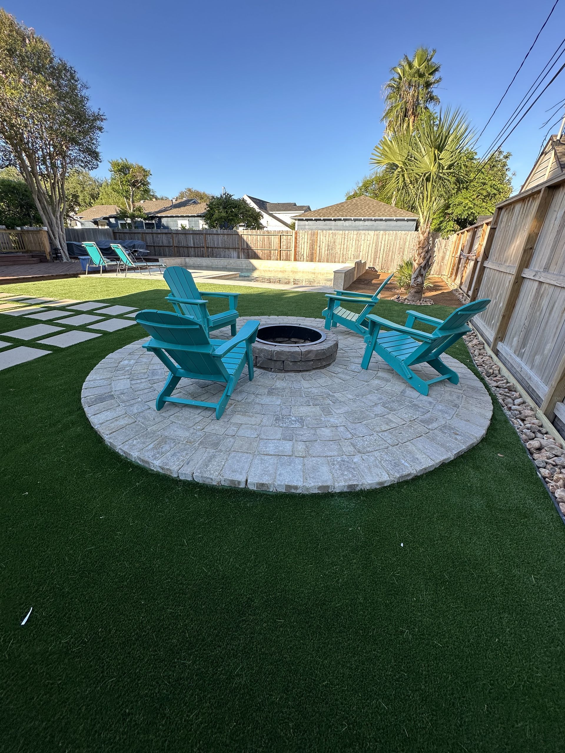 Three turquoise Adirondack chairs arranged around a circular stone fire pit in a backyard with artificial grass.