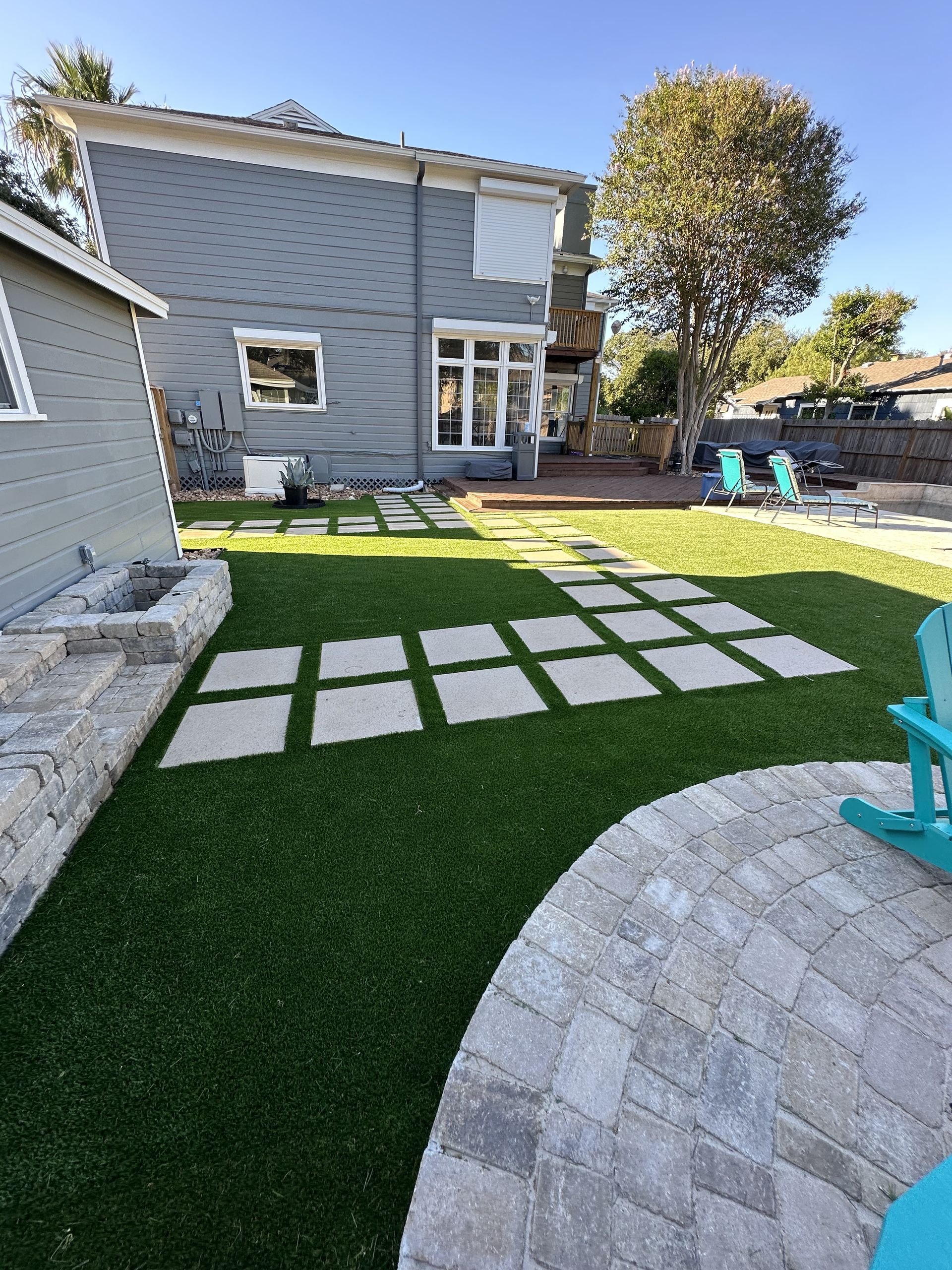 A backyard with a green lawn, stone paver path, patio, and a two-story gray house under a clear blue sky.