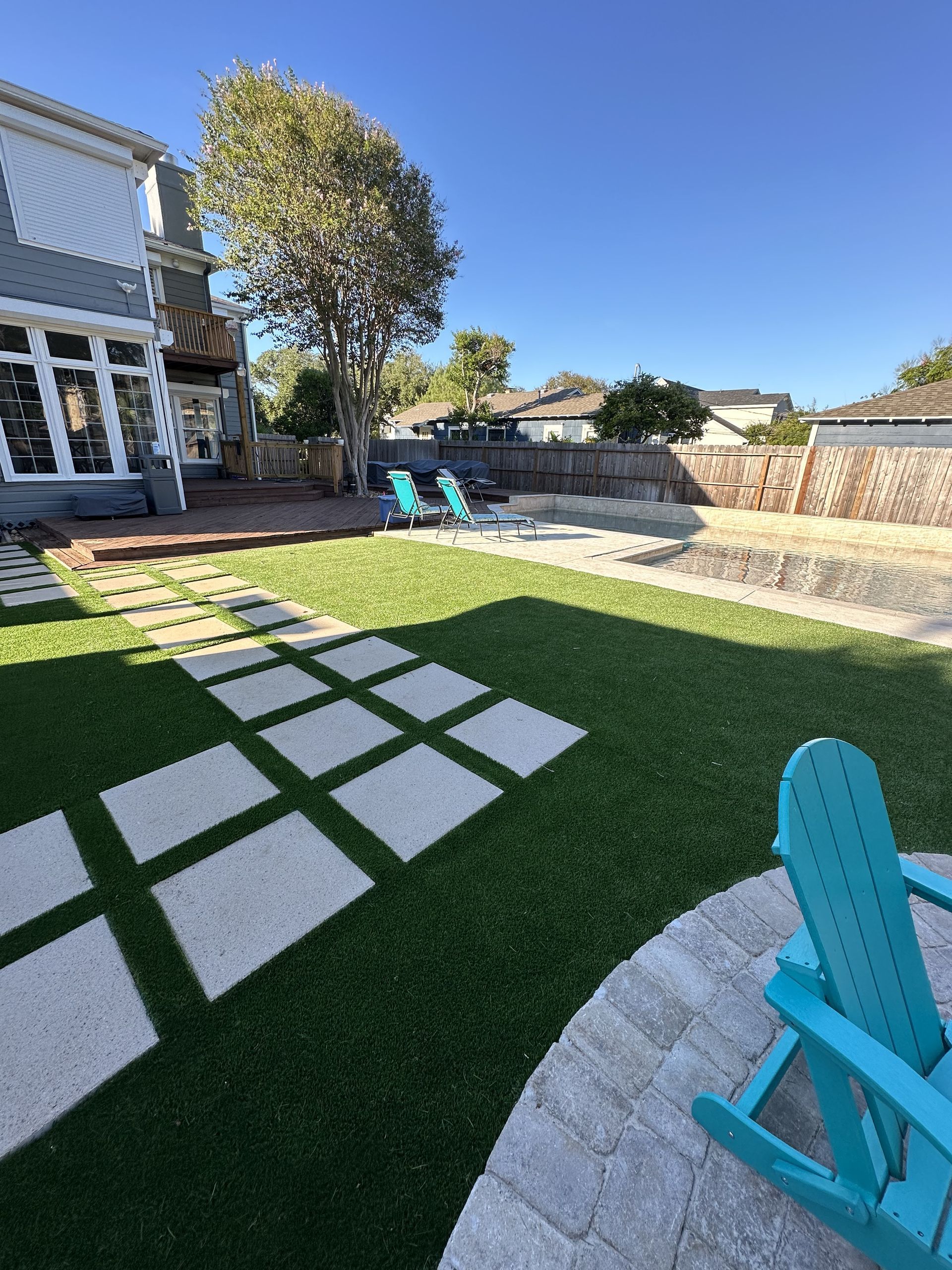 A backyard with a green lawn, stone paver path, and teal Adirondack chairs on a stone patio under a clear blue sky.