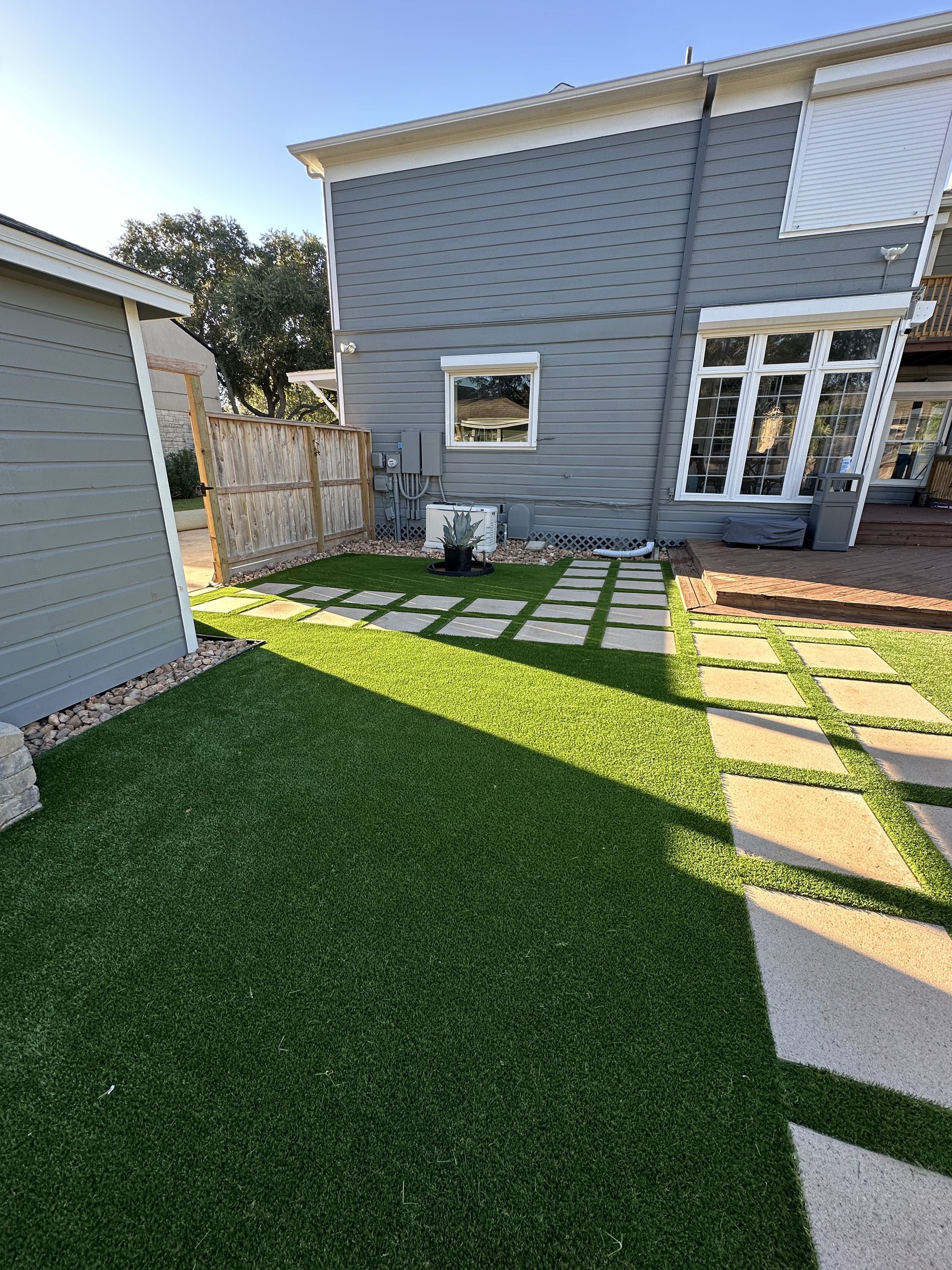 A backyard features a manicured green lawn with light gray paver stones arranged as a path leading toward a gray house.