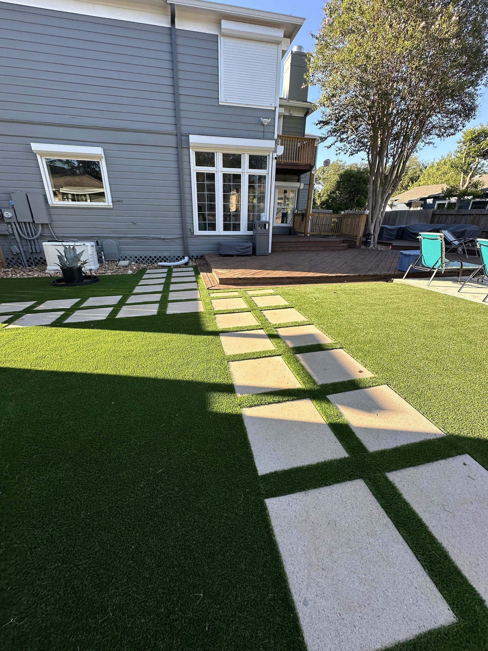 A backyard with a green lawn, stone paver path, and a grey house exterior under a clear blue sky.