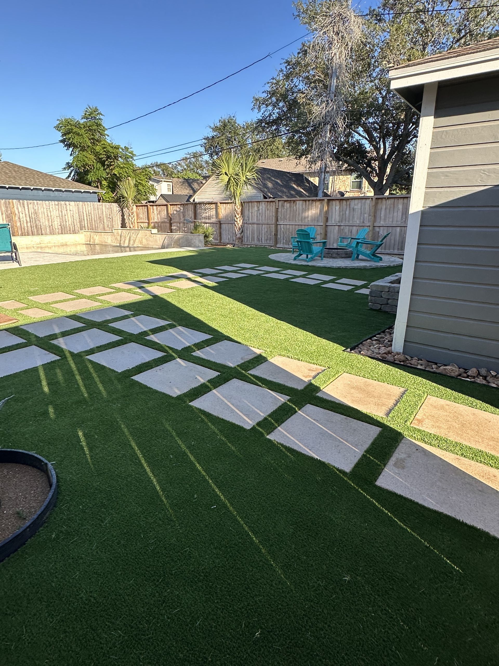 A backyard with artificial grass, square paver paths, and two blue Adirondack chairs near a corner fire pit.